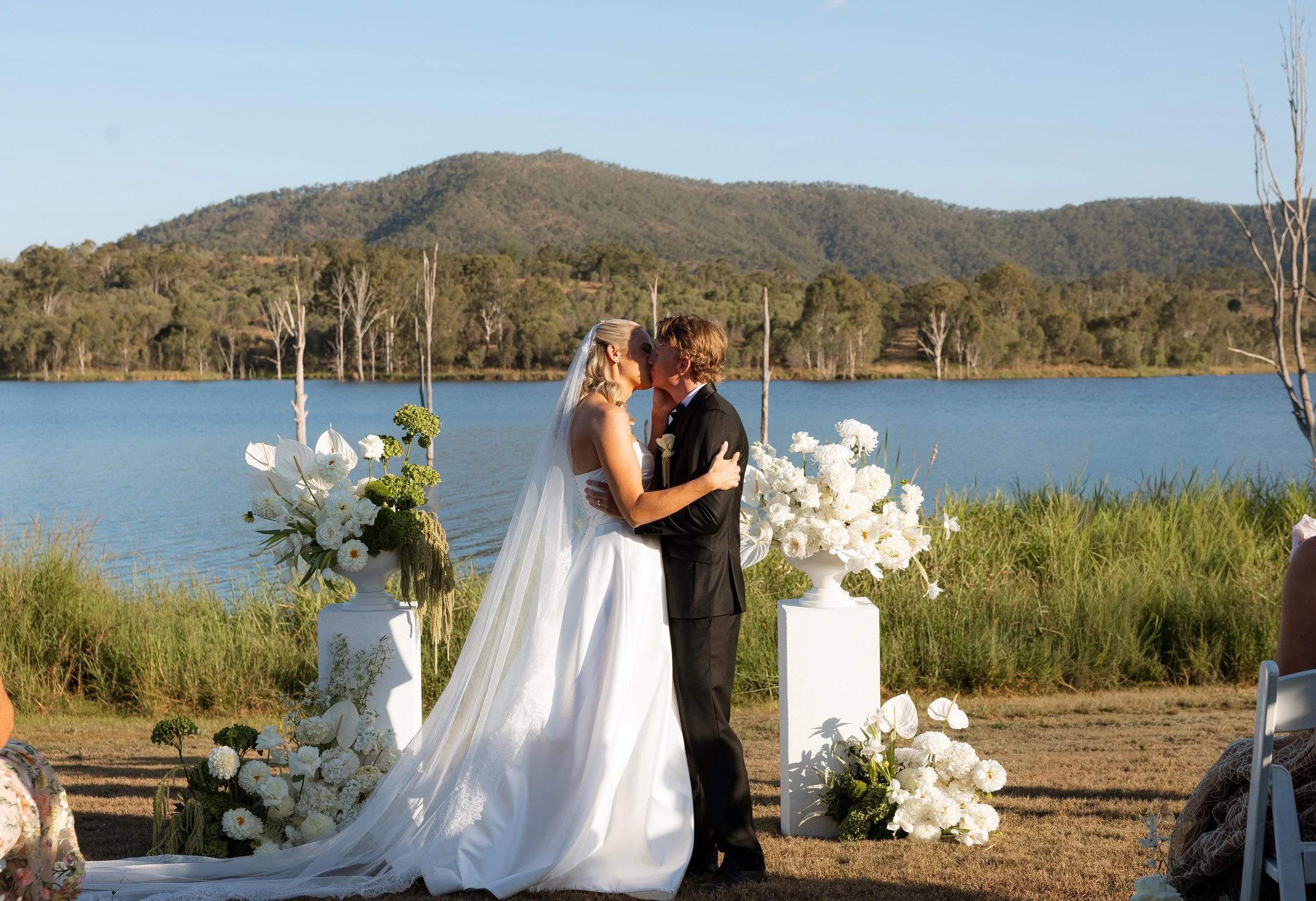 A bride and groom sharing a kiss at an outdoor wedding ceremony by a lake with mountains in the background, decorated with white flowers and greenery.