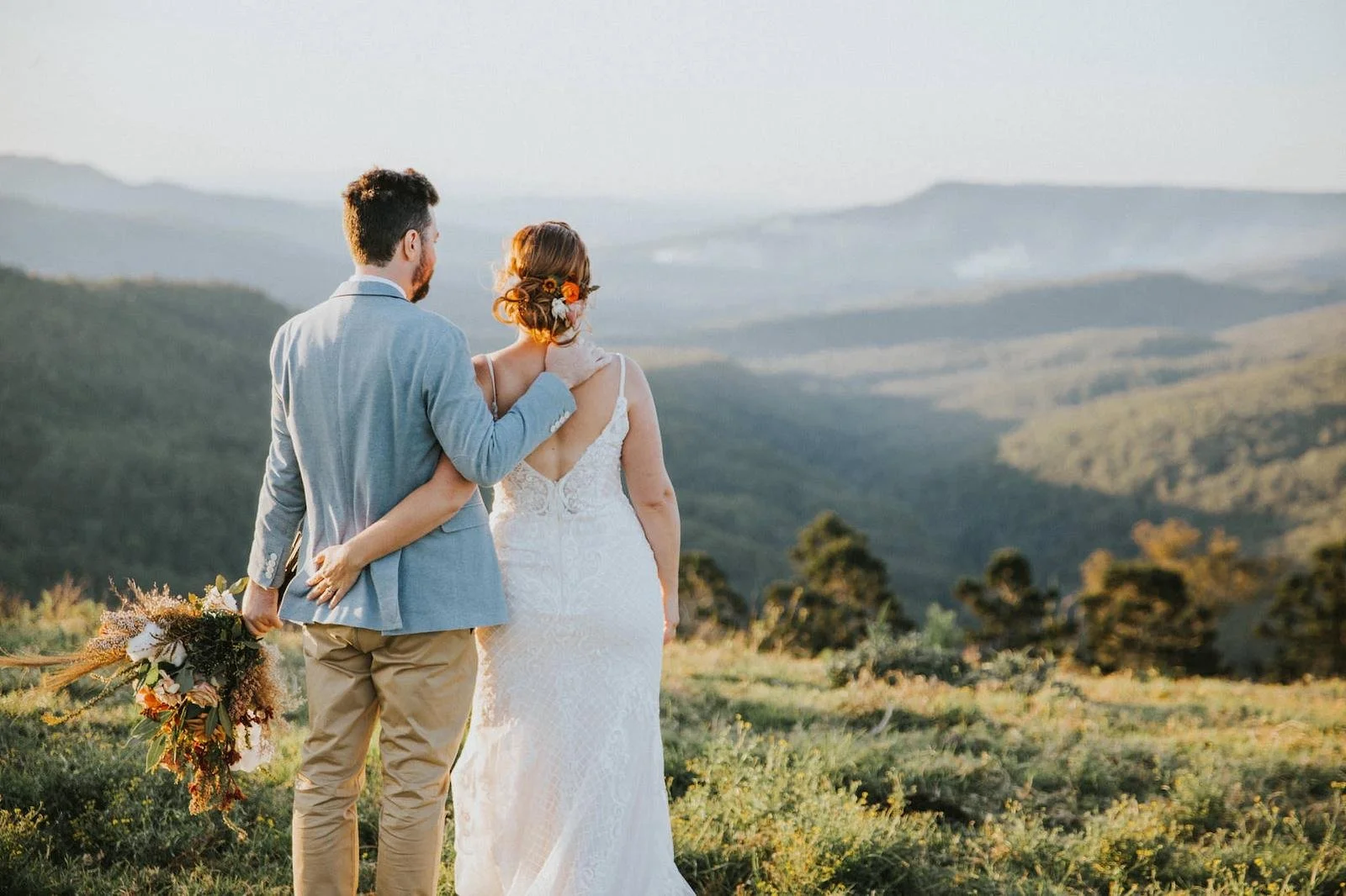 A newlywed couple stands outdoors in a lush green field, overlooking rolling hills and mountains, during sunset. The groom is wearing a light blue blazer and beige pants, with his arm around the bride. The bride is wearing a white lace wedding dress,