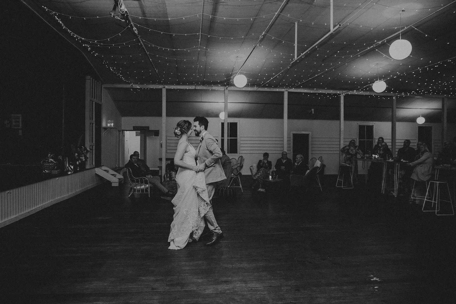 A black and white photo of a bride and groom dancing in a large hall decorated with string lights and hanging lanterns. The bride is wearing a lace wedding gown and the groom is in a suit. Guests are seated at tables in the background, watching the c