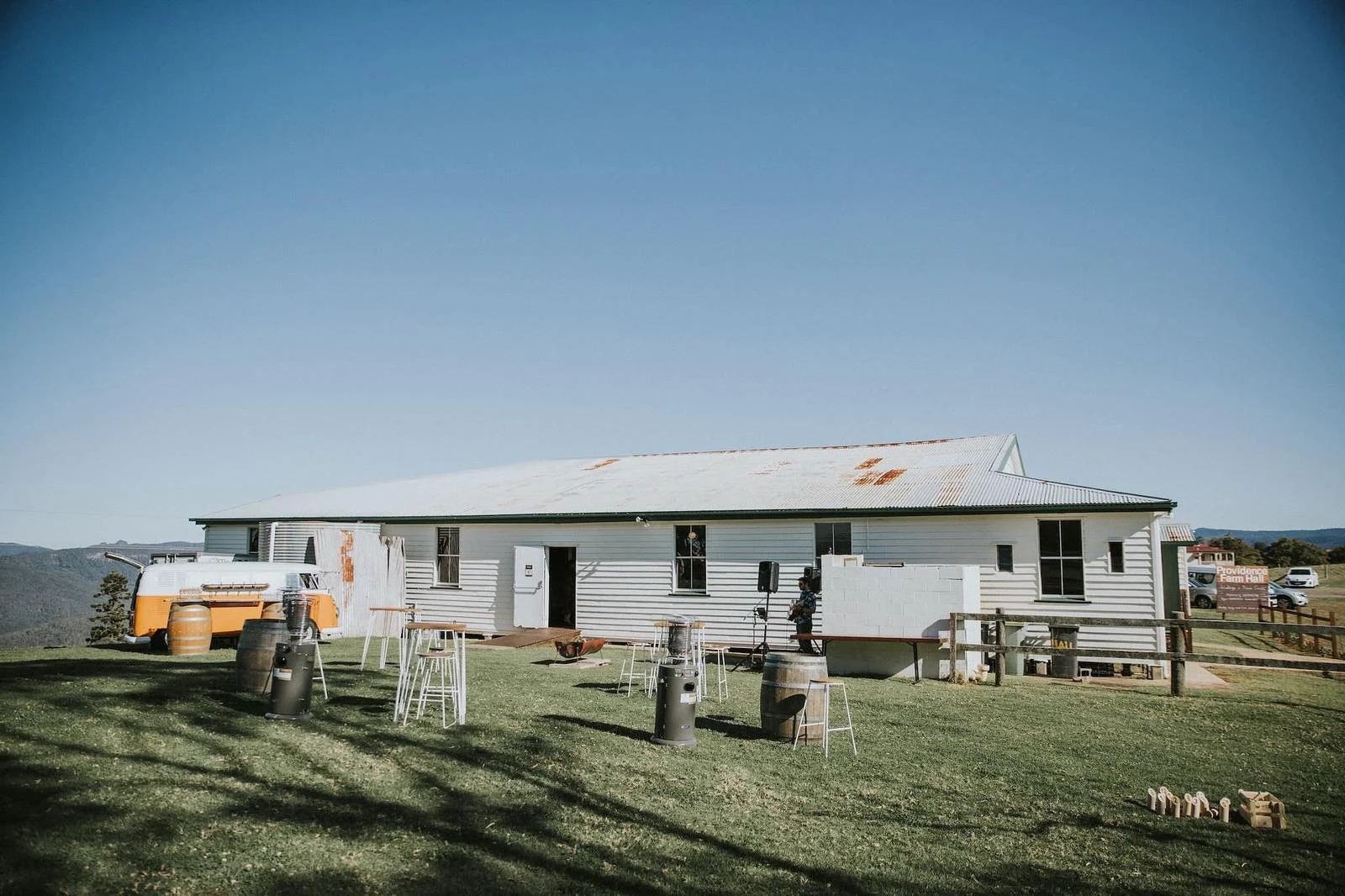 A white, rustic building with a metal roof shows signs of rust. There are several windows, and an open door in the center. Outside, there are barrels, tables, stools, and metal heaters on a grassy lawn. A vintage camper trailer is parked to the left,