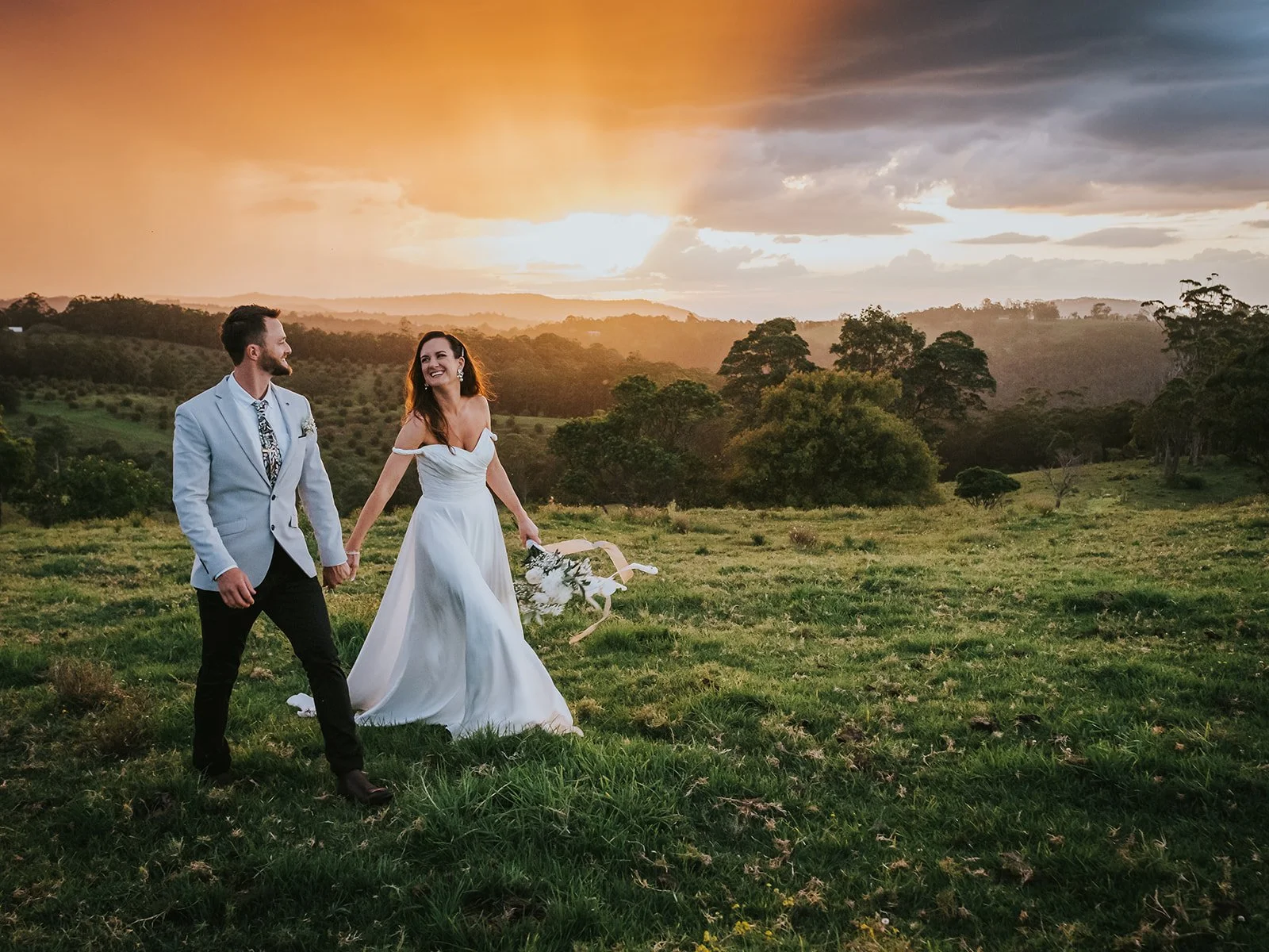 A newlywed couple holding hands and smiling at each other in a grassy field at sunset, with a scenic background of trees, rolling hills, and a dramatic sky.