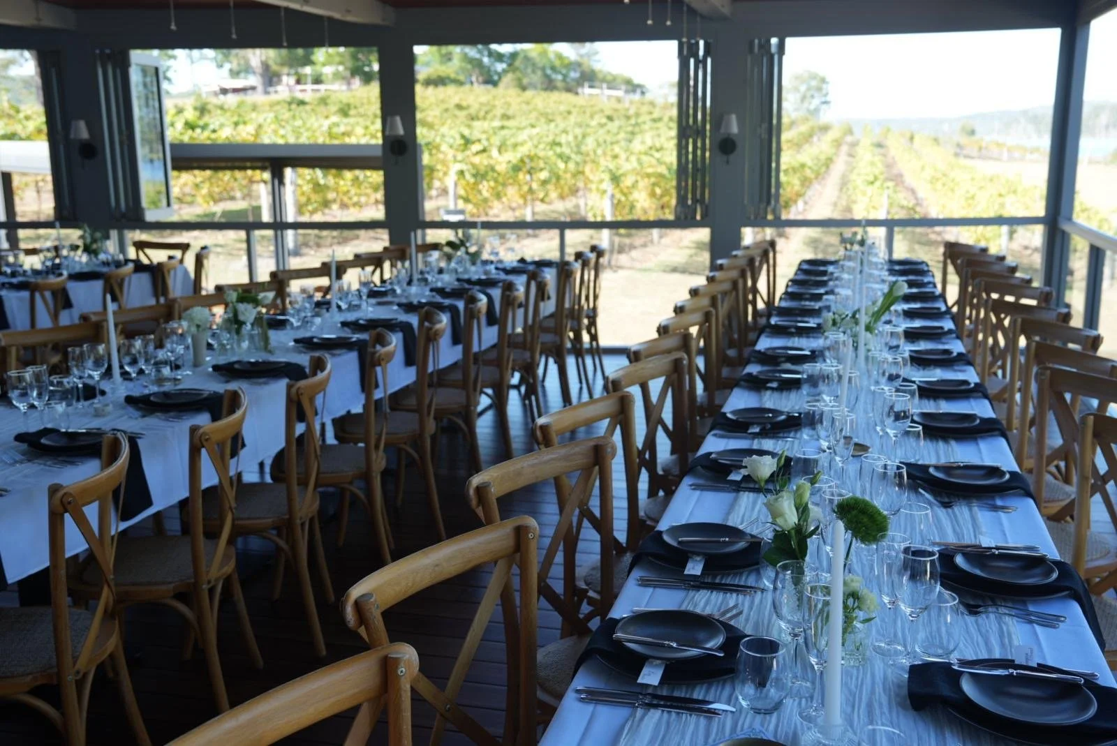 Indoor event space with long dining tables set for a formal occasion, decorated with white tablecloths, black plates, wine glasses, and floral centerpieces, with large windows overlooking a vineyard or farm landscape.