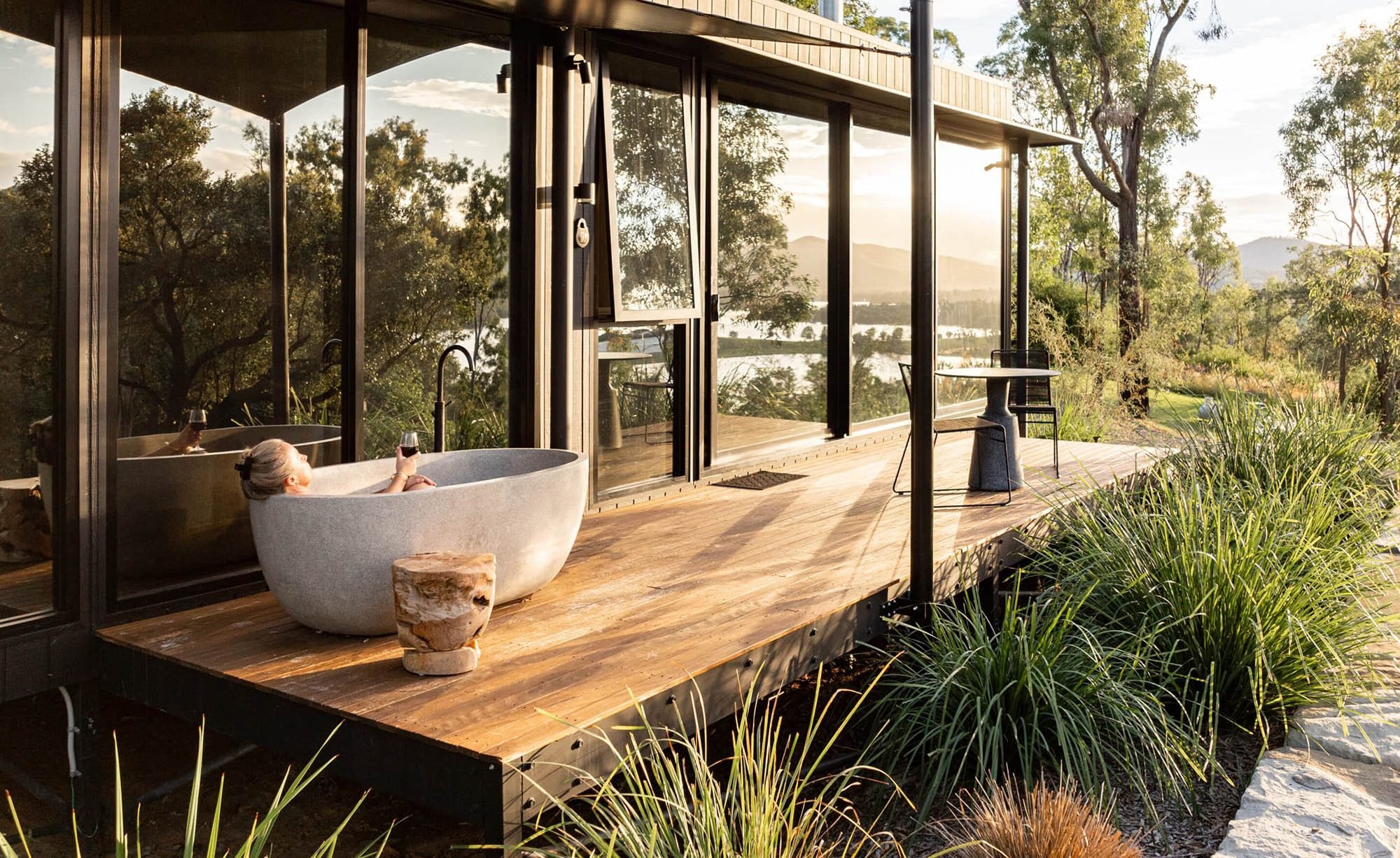 A woman relaxing in a round freestanding bathtub on a wooden deck, holding a glass of wine, with a scenic view of trees, water, and distant mountains through large glass doors and windows.