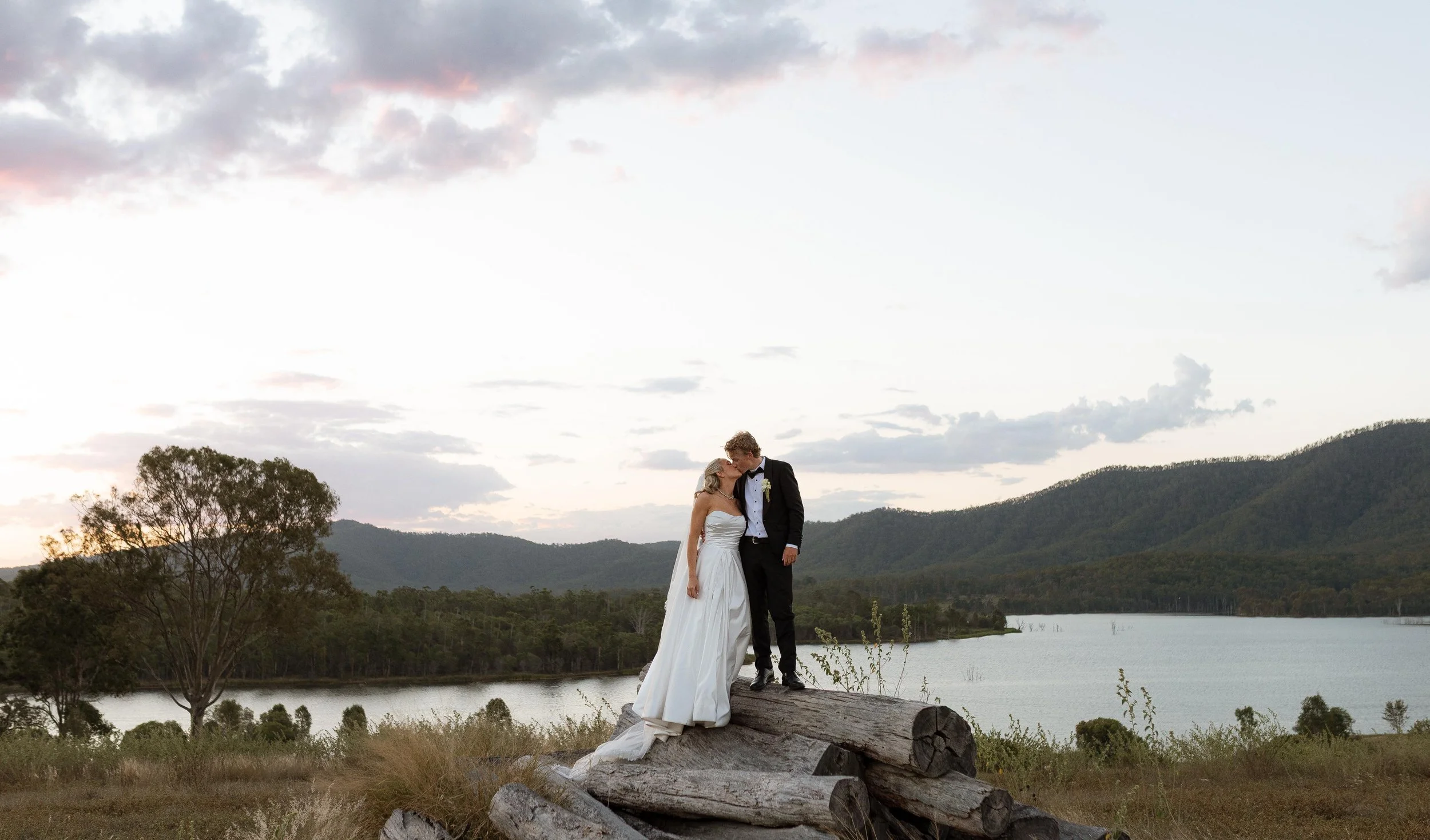 A bride and groom sharing a kiss outdoors on a large fallen log, with a scenic backdrop of trees, water, hills, and a cloudy sky at sunset.