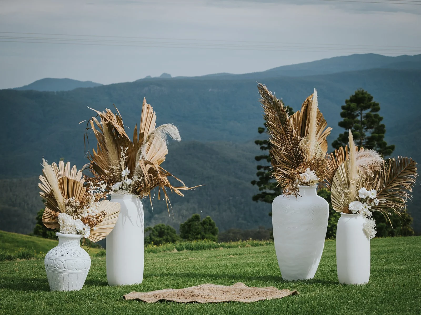Four white vases filled with dried palm leaves, pampas grass, and white flowers placed on green grass with mountains and trees in the background.