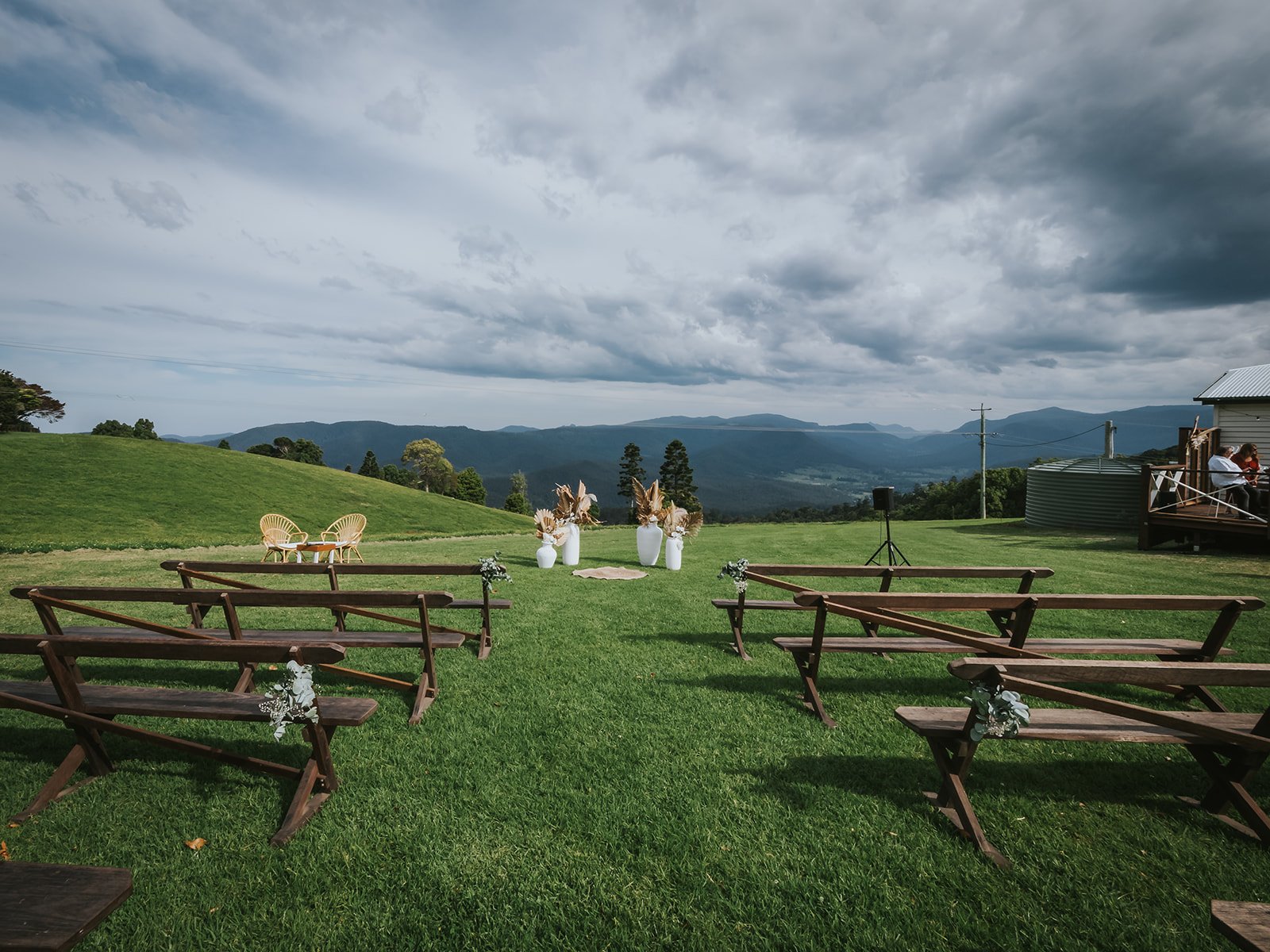 Outdoor wedding ceremony setup with wooden benches on green grass, white vases with dried flowers, two wicker chairs, and scenic mountain view under cloudy sky.