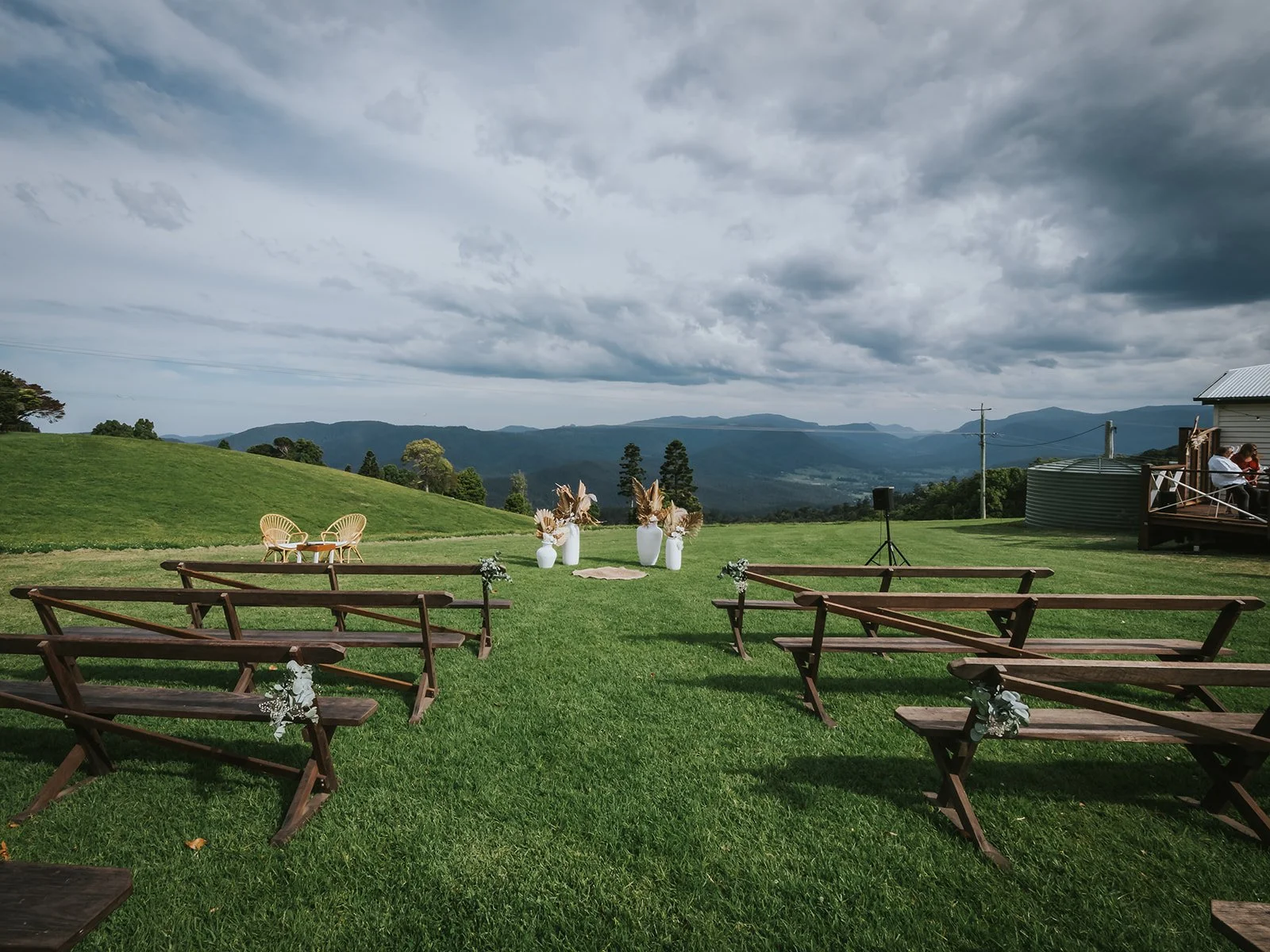 Outdoor wedding setup on a lush green field with mountain views, wooden benches with small flower arrangements, large white vases with dried plants, and a cloudy sky overhead.