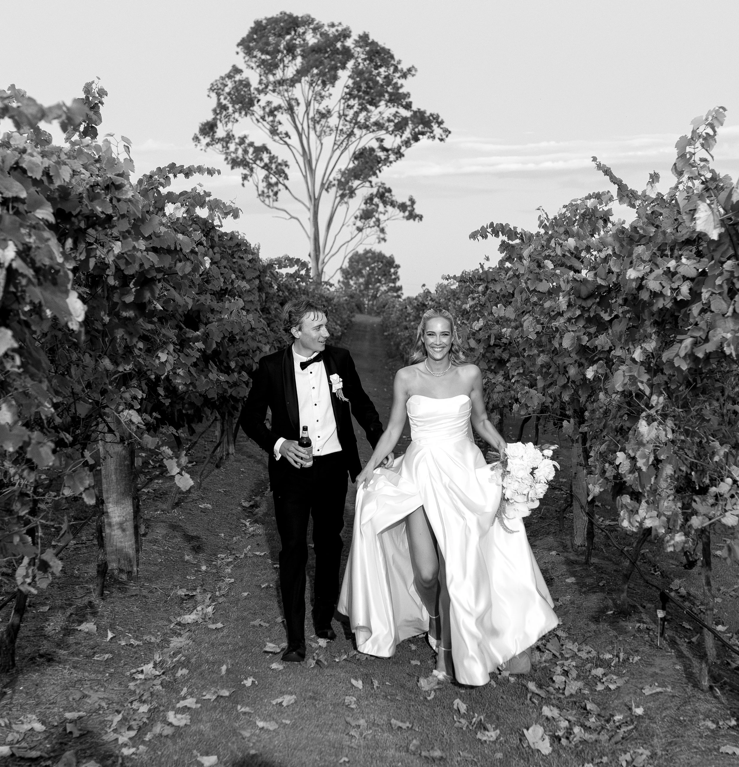 Black and white wedding photograph of a smiling bride and groom walking hand in hand through a vineyard, dressed in wedding attire, with grapevines on either side and a tall tree in the background.