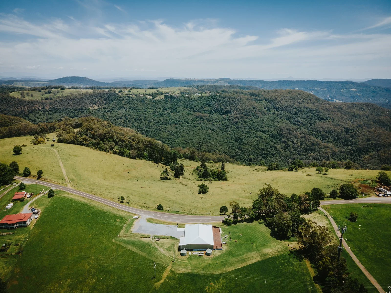 Aerial view of green rolling hills, with a few buildings and a winding road in a rural area under a partly cloudy sky.