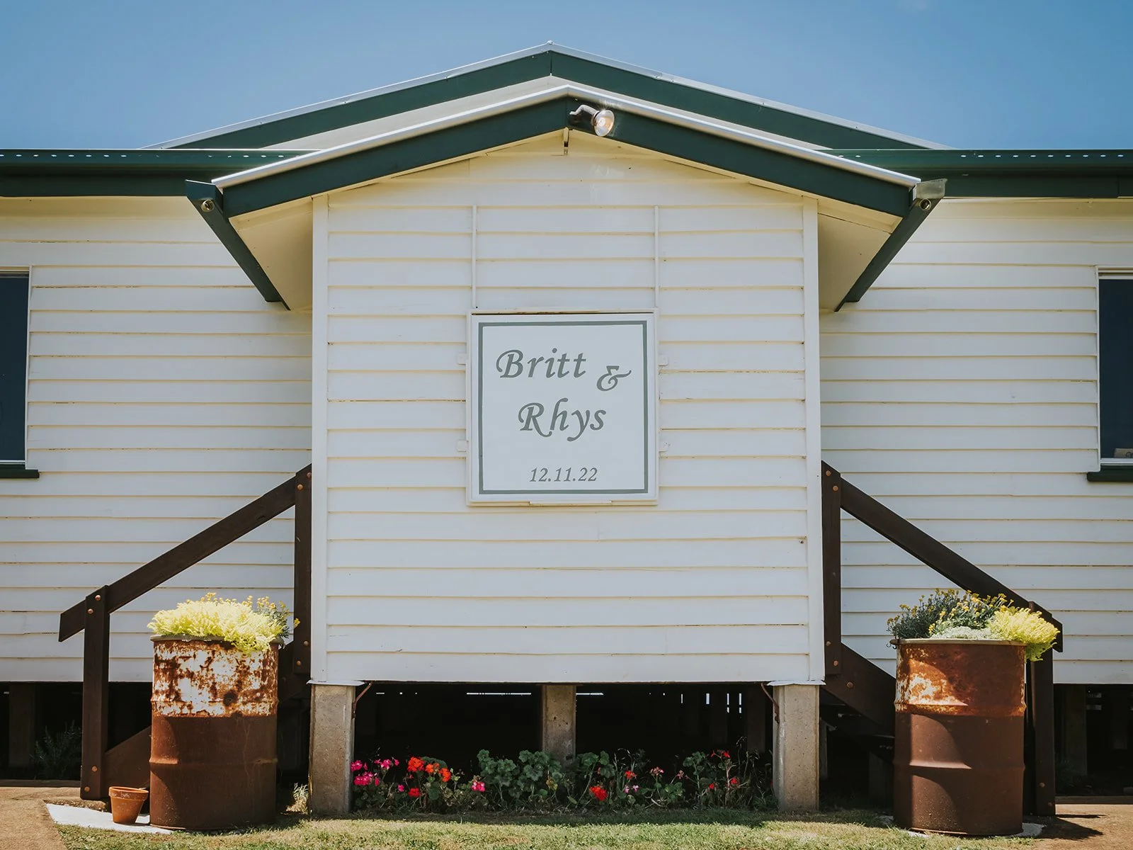 A white house with a gable roof and a sign in the center that reads 'Britt & Rhys 12.11.22'. There are two large rusted planters with yellow and white flowers on either side of the steps in front of the house. Small colorful flowers are visible benea