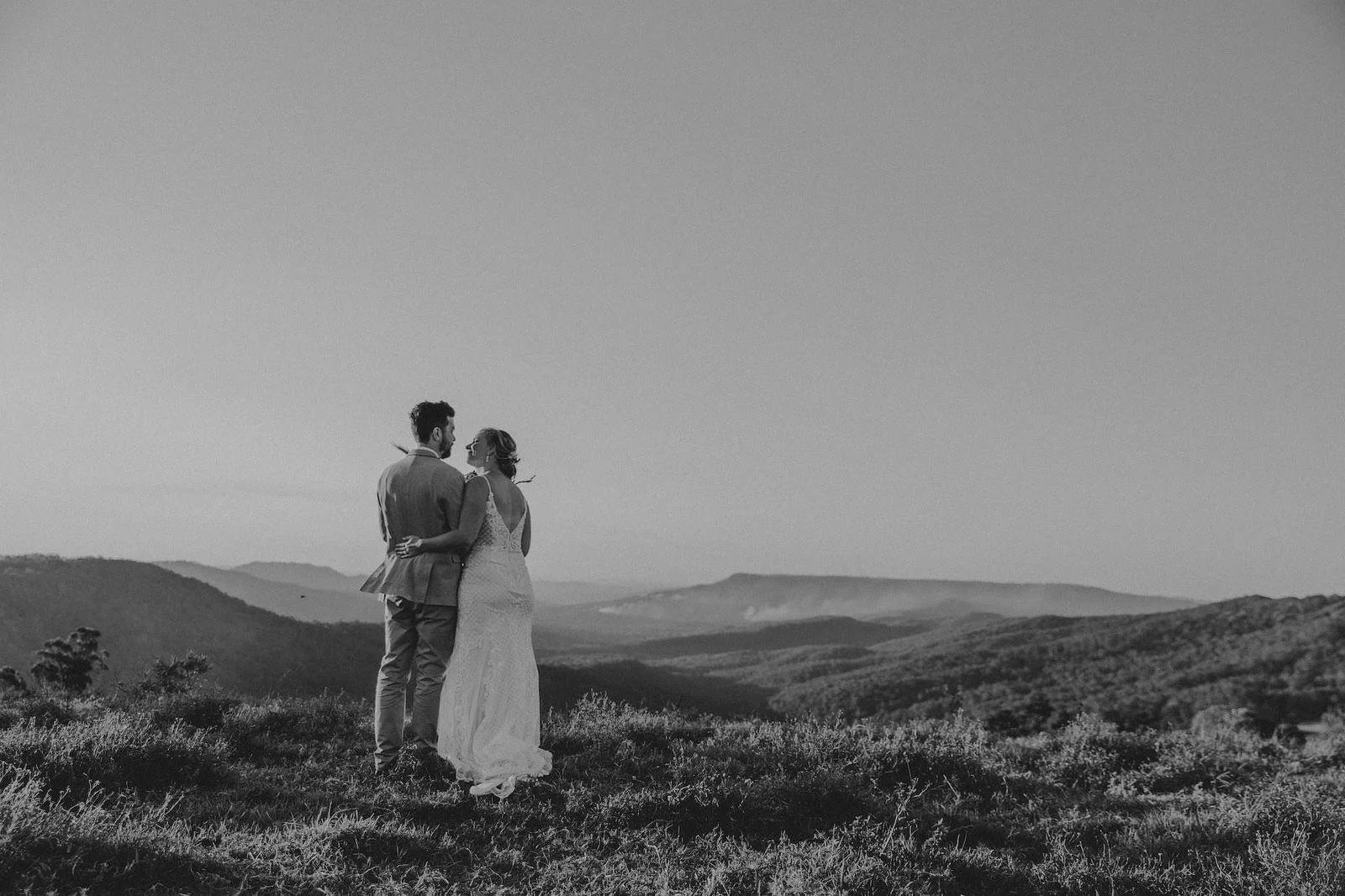 Black and white photo of a newlywed couple standing in a grassy field, embracing and looking at each other with mountains in the background.