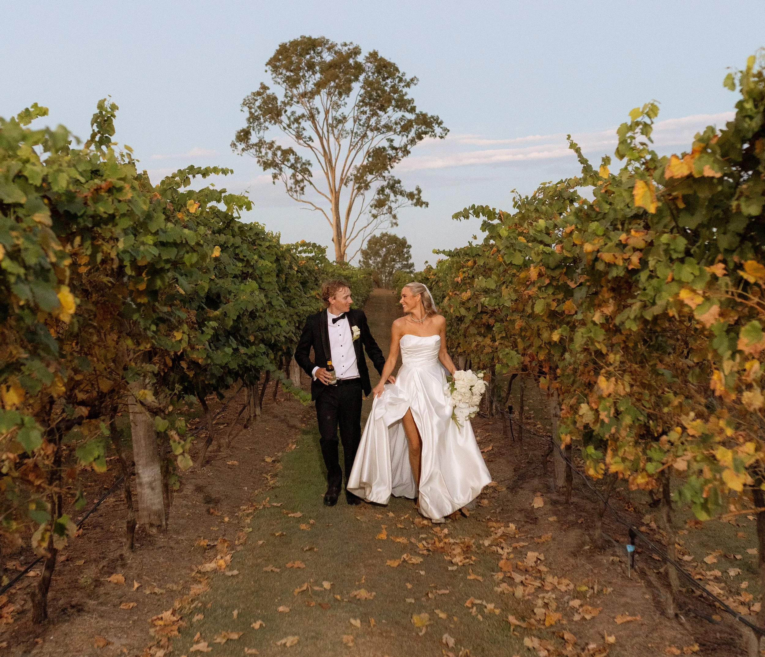 A bride and groom walking hand-in-hand through vineyard rows during sunset, with the groom in a tuxedo and the bride in a strapless wedding gown holding a bouquet of white flowers, surrounded by autumn-colored leaves.