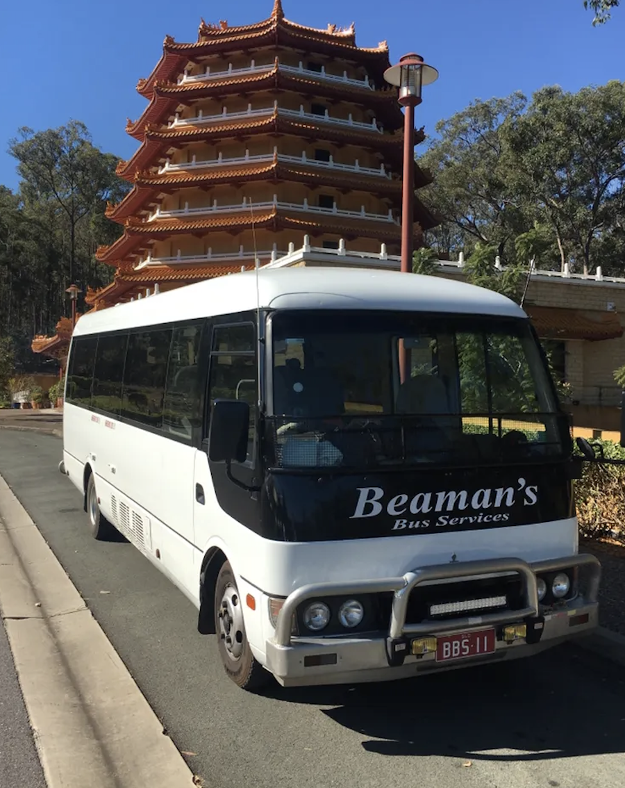 A white bus labeled 'Beamans Bus Services' parked on the side of the road in front of a traditional multi-tiered Asian pagoda with orange and white roofs, surrounded by green trees, under a clear blue sky.