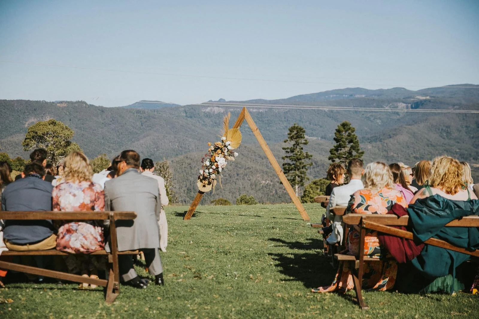 Outdoor wedding ceremony setup with wooden benches, floral decoration, and an arch against a mountain landscape.