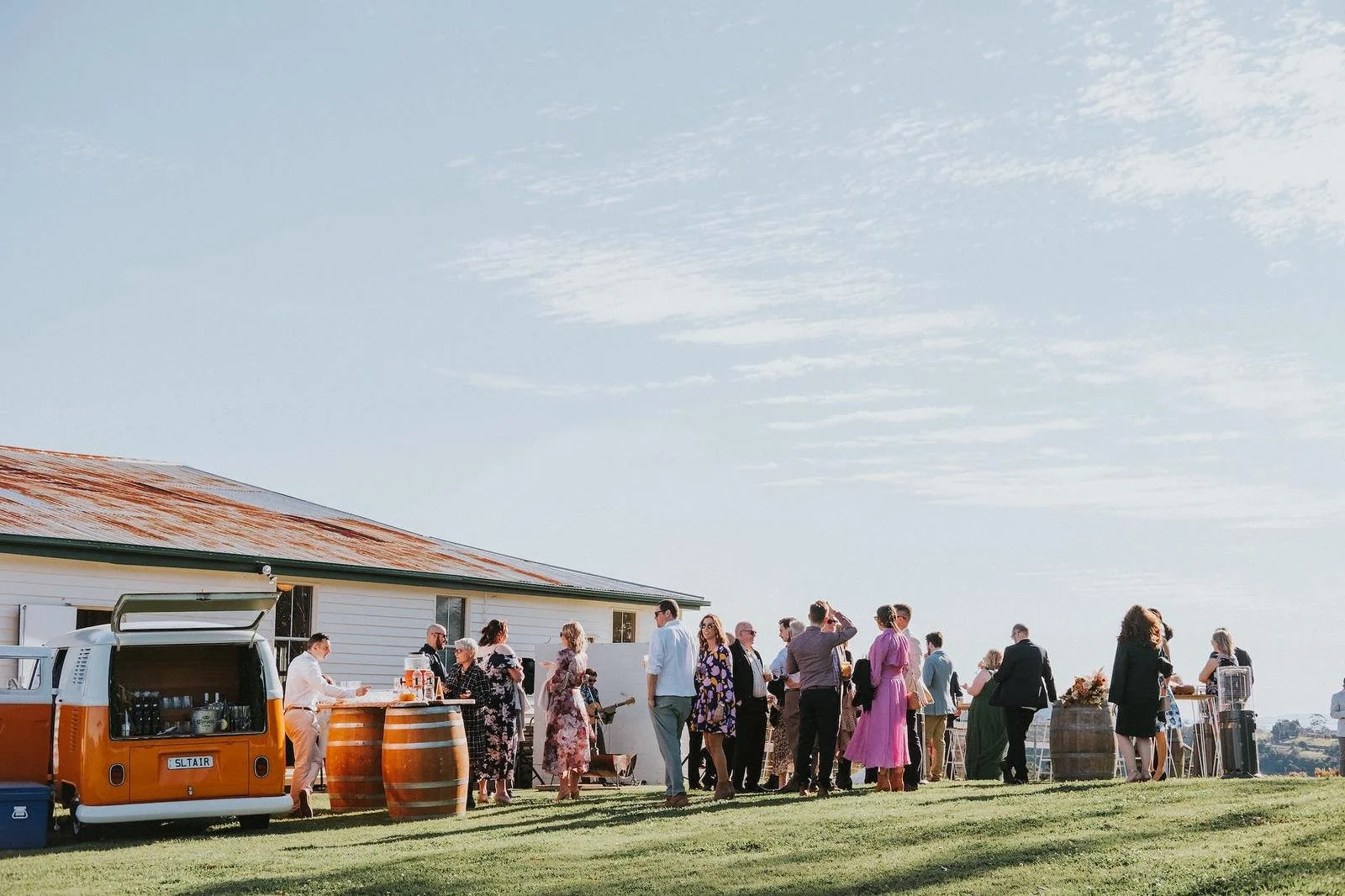 Outdoor gathering of people at a daytime event, with a vintage orange and white van, barrel tables, and a white building, set against a partly cloudy sky.