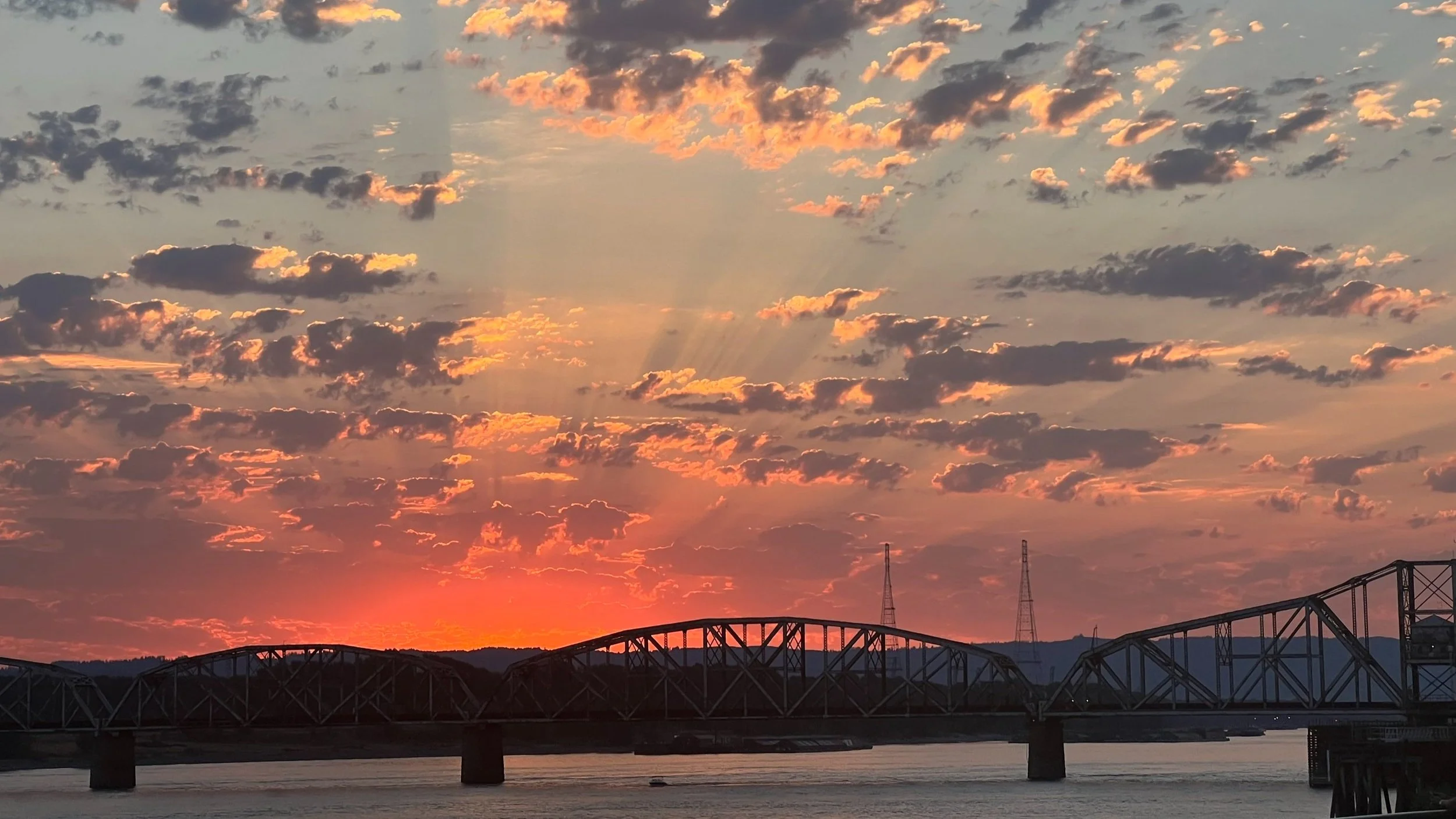 EDP DESIGN PHOTOGRAPHY Sunset over a river with a silhouetted bridge and scattered clouds, emitting rays of light.