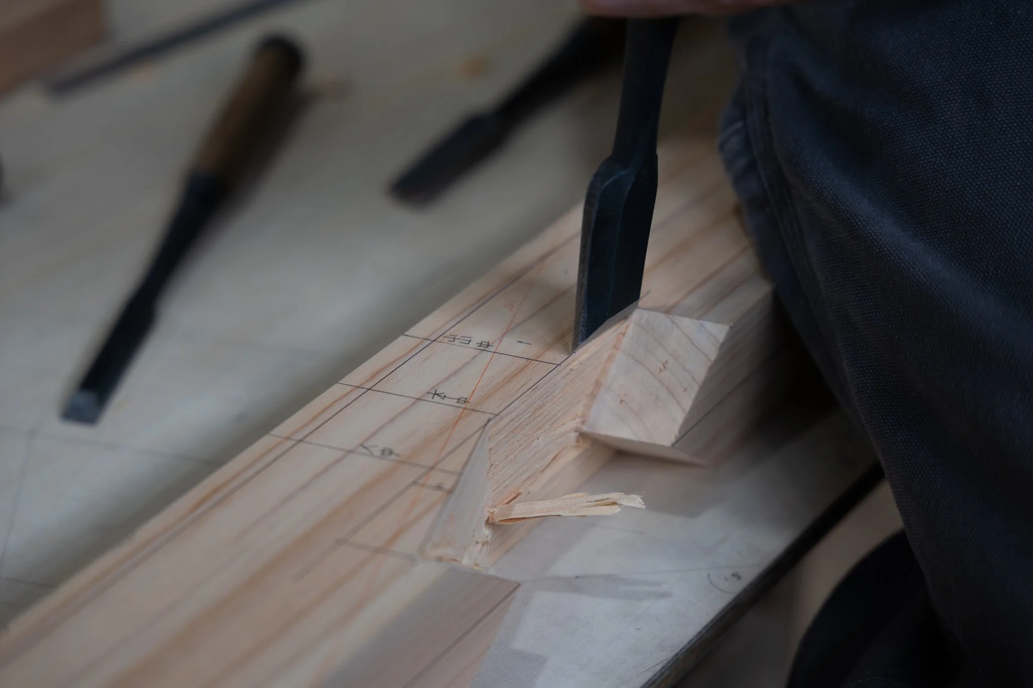 Close-up of a woodworking project with marked measurements on a piece of wood, black tools, and a person's arm in a black sleeve.