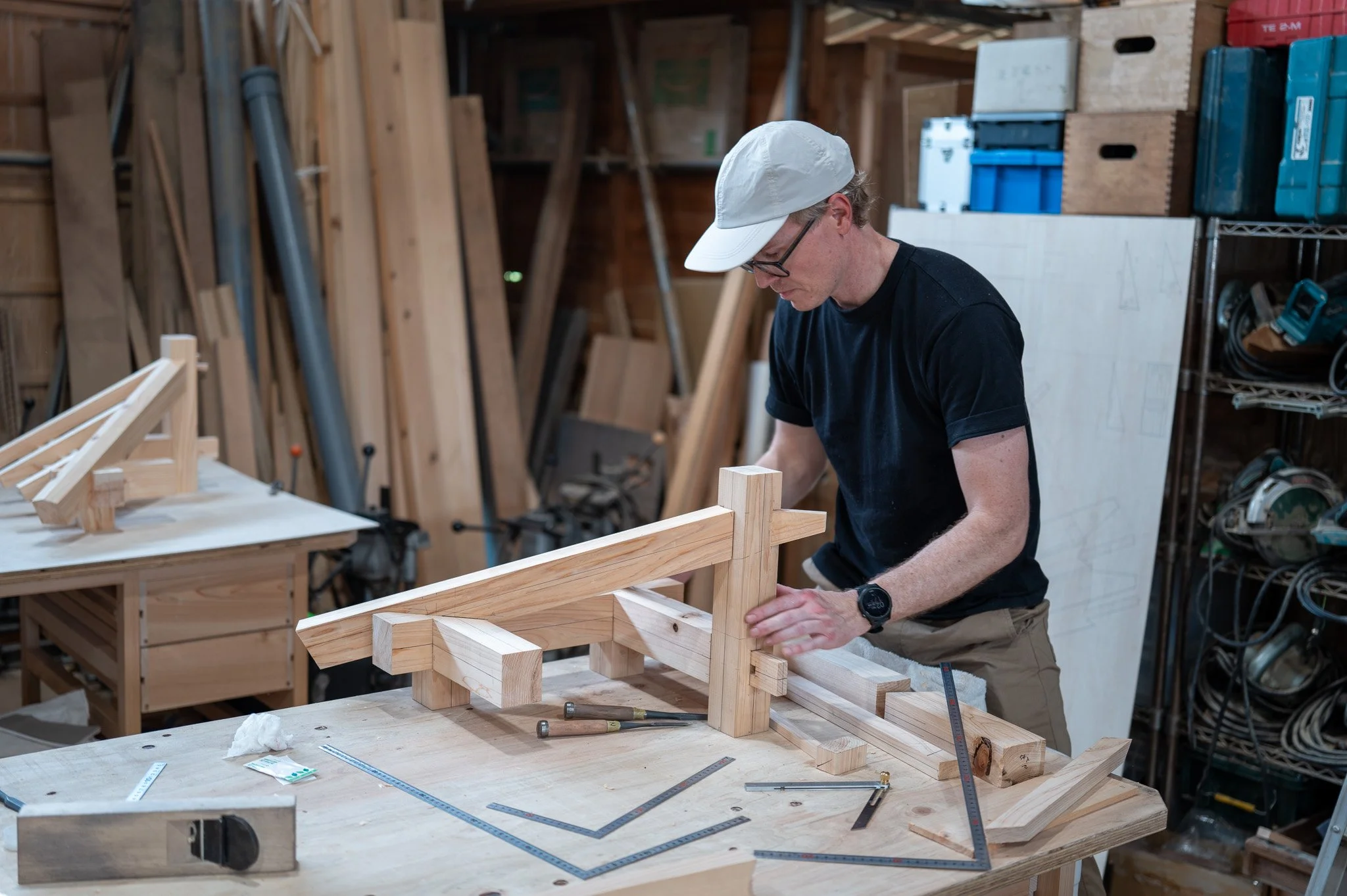 A man in a white cap, glasses, and black shirt works on a woodworking project in a cluttered workshop with wood and tools. He appears to be measuring or assembling a wooden structure.