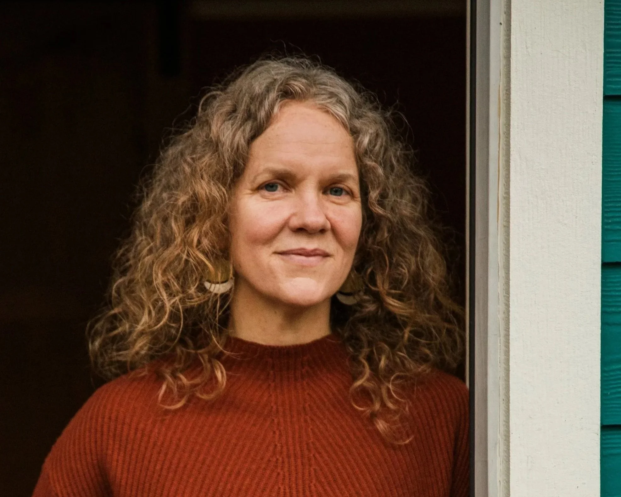 A woman with curly hair, wearing a rust-colored top, standing outdoors near a building with teal and white siding.