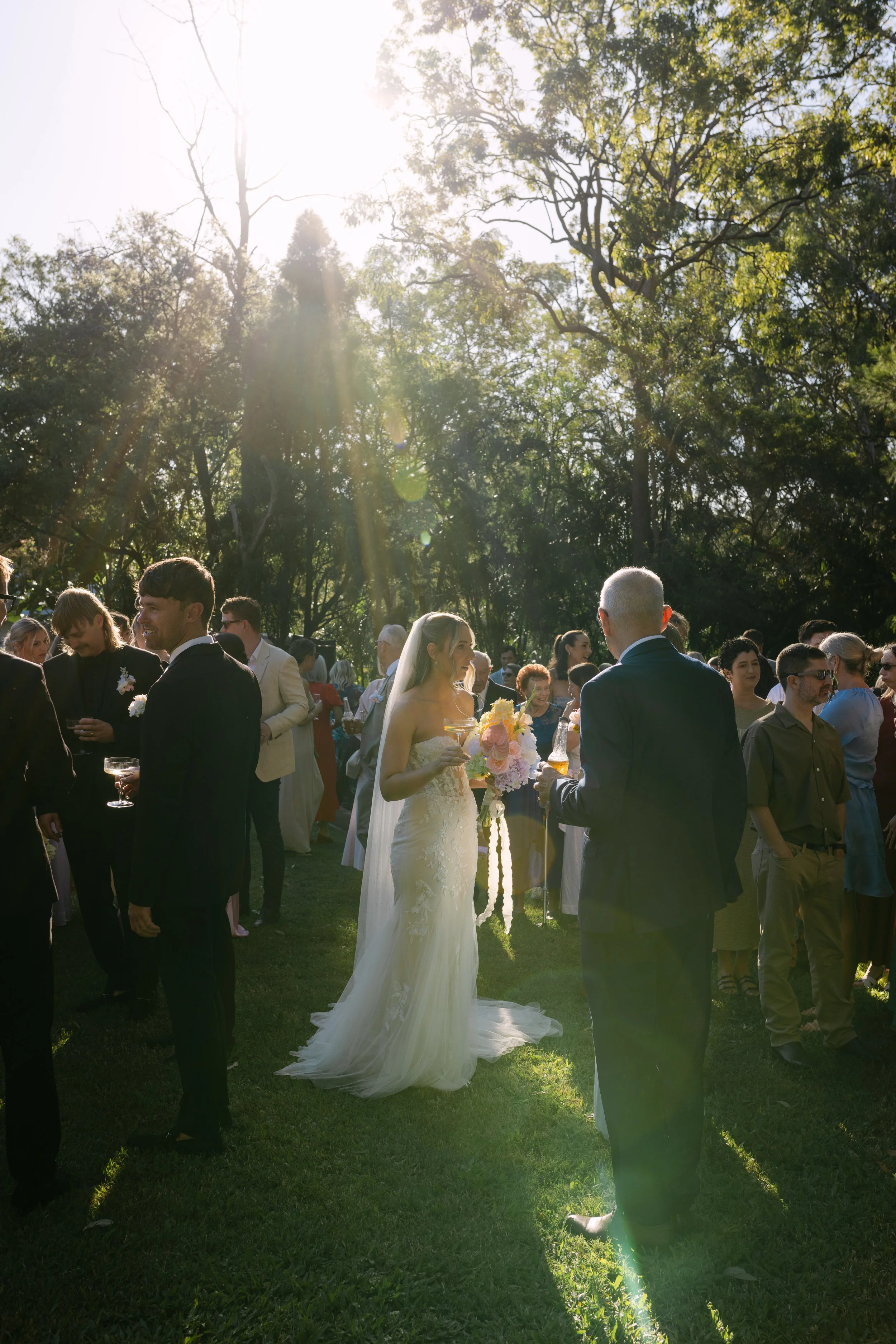 A wedding gathering outdoors with guests mingling on a grassy area under trees, sunlight shining through the trees, with a bride in a white gown and veil holding a bouquet, and men in suits.