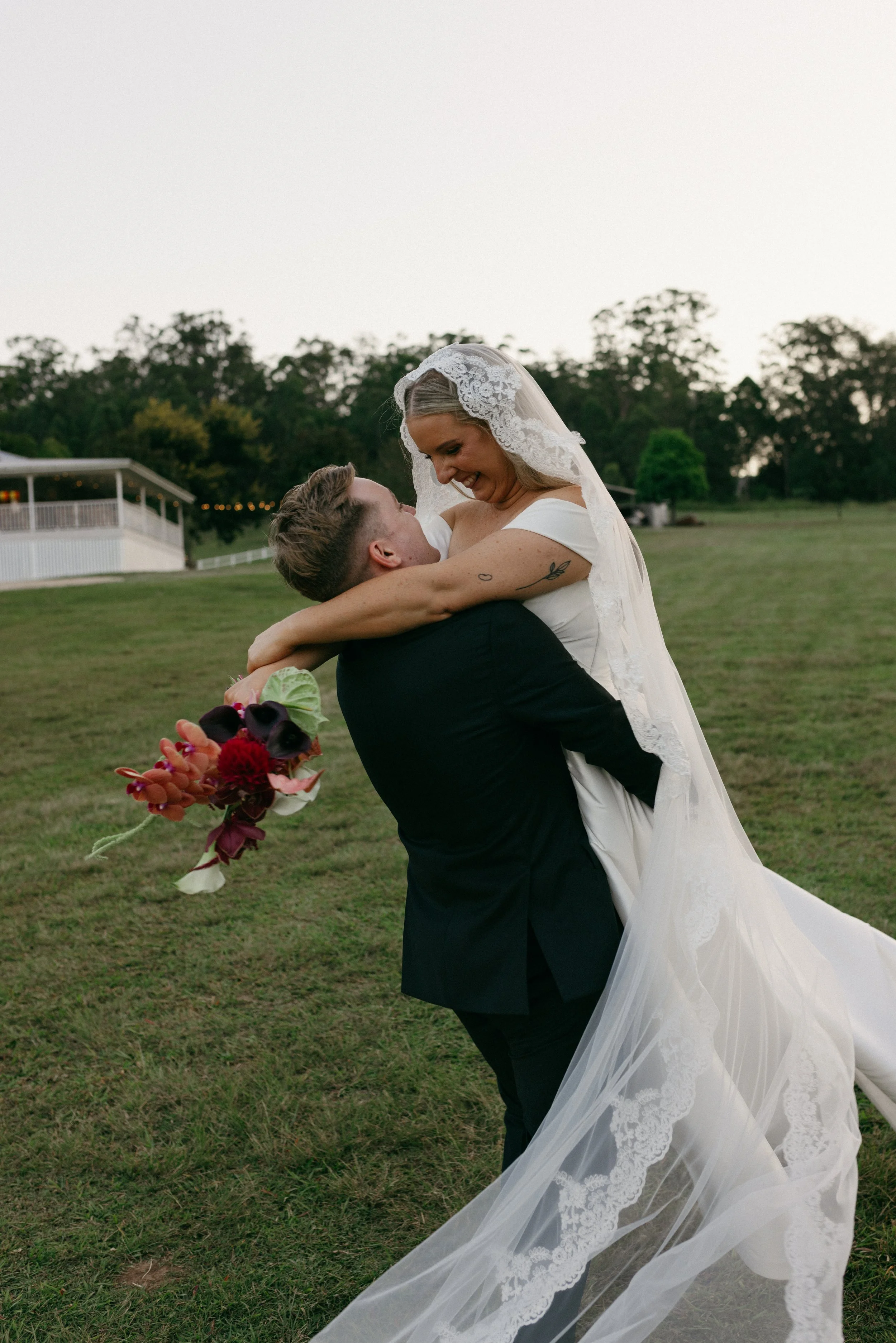 A bride and groom on a grassy field during sunset, with the groom holding the bride in his arms, and the bride smiling and looking down at him. The bride is wearing a white wedding dress with lace details and a veil, and holds a bouquet of dark red, 