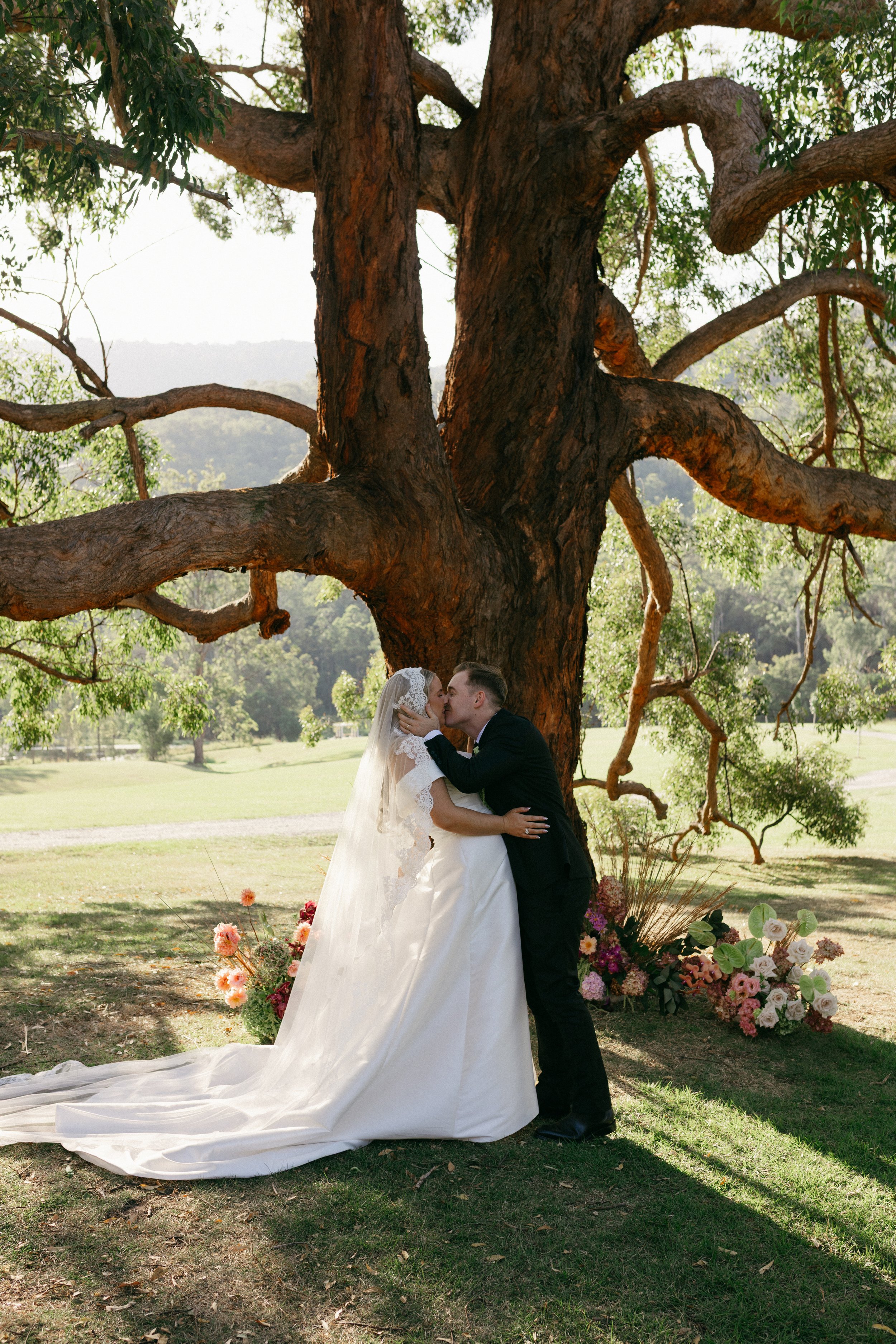 A bride and groom kiss during their outdoor wedding ceremony beneath a large tree, with floral arrangements on the ground.
