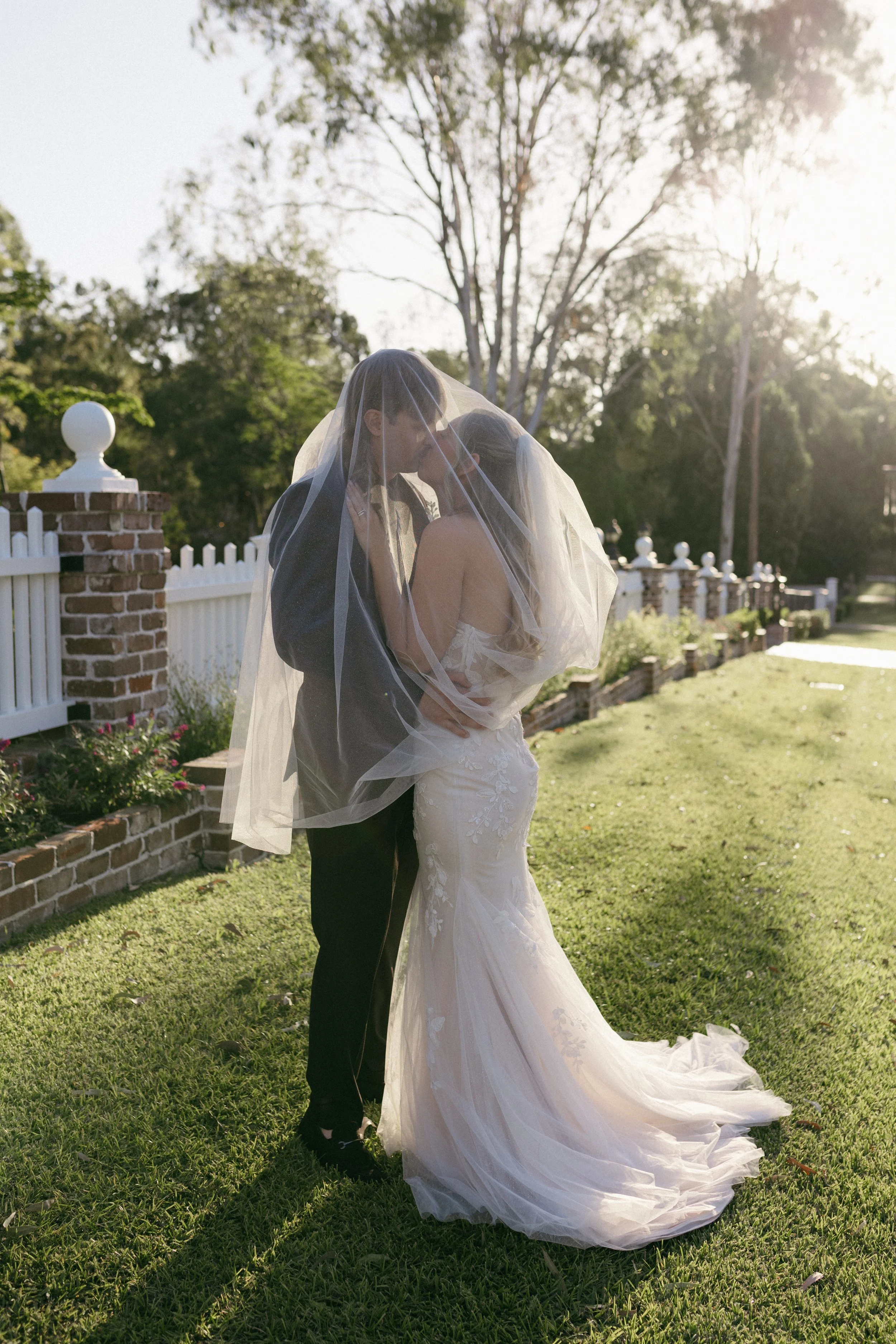 A bride and groom share a kiss outdoors during sunset, with the bride wearing a white wedding gown and veil, and the groom in a black suit, in front of a white fence and trees.