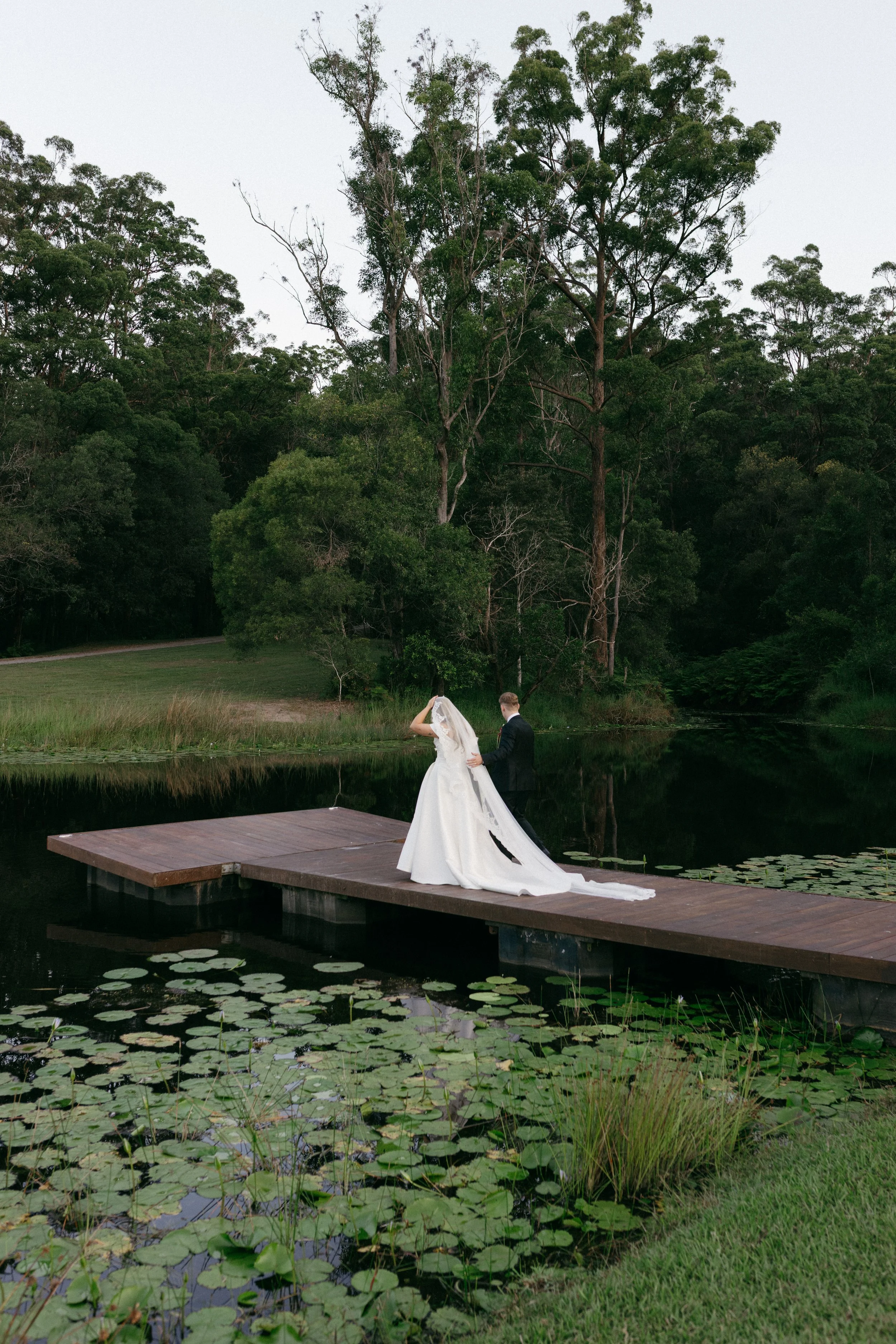A bride and groom stand on a wooden dock by a pond surrounded by greenery and tall trees, with lily pads on the water. The bride is wearing a long white wedding gown with a veil, and the groom is in a black suit. They are holding hands and facing eac