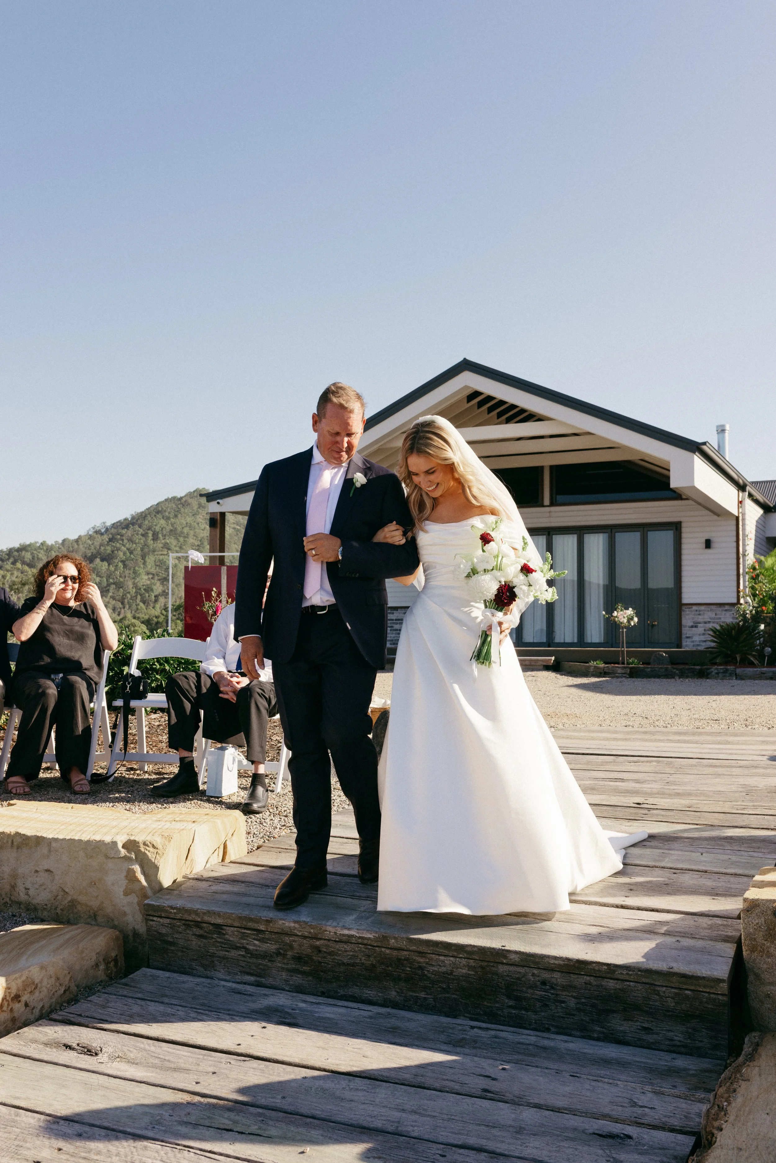 A bride in a white wedding gown holding a bouquet, walking down a wooden path while being assisted by a man in a dark suit at an outdoor wedding ceremony. Guests are seated and watching, with a house and mountains in the background under a clear sky.