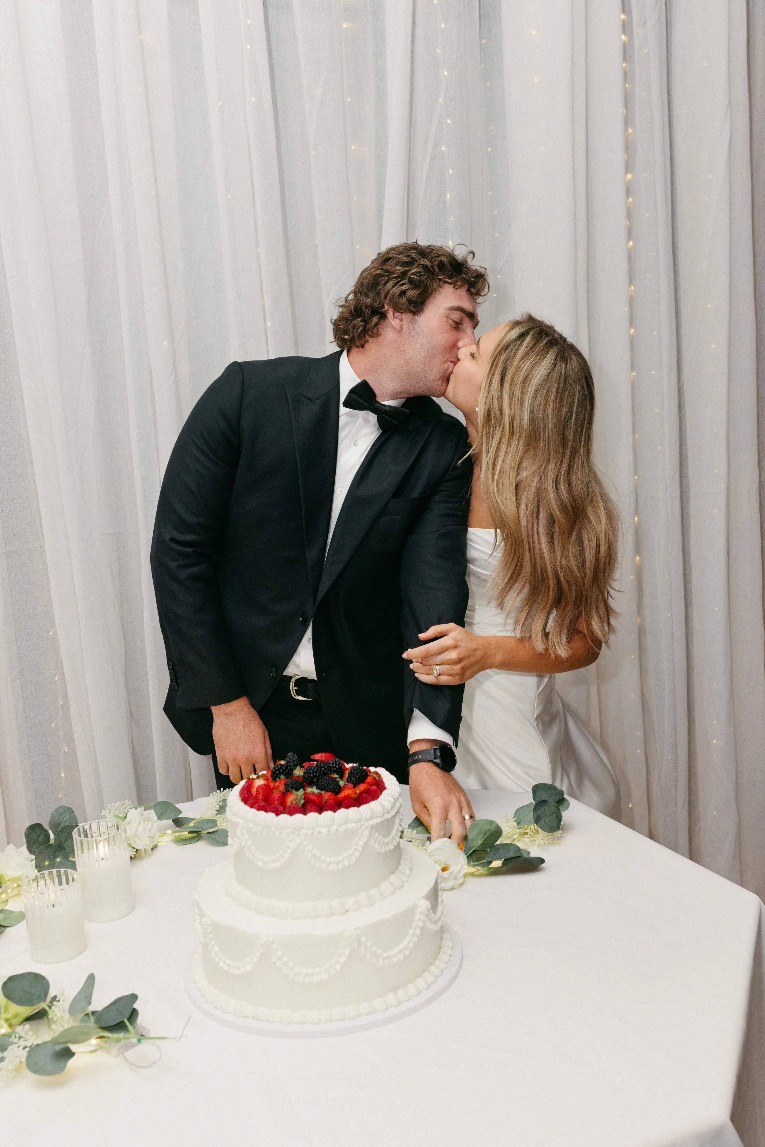A newlywed couple sharing a kiss at their wedding reception, standing in front of a wedding cake decorated with berries and greenery, with a decorated backdrop and fairy lights in the background.