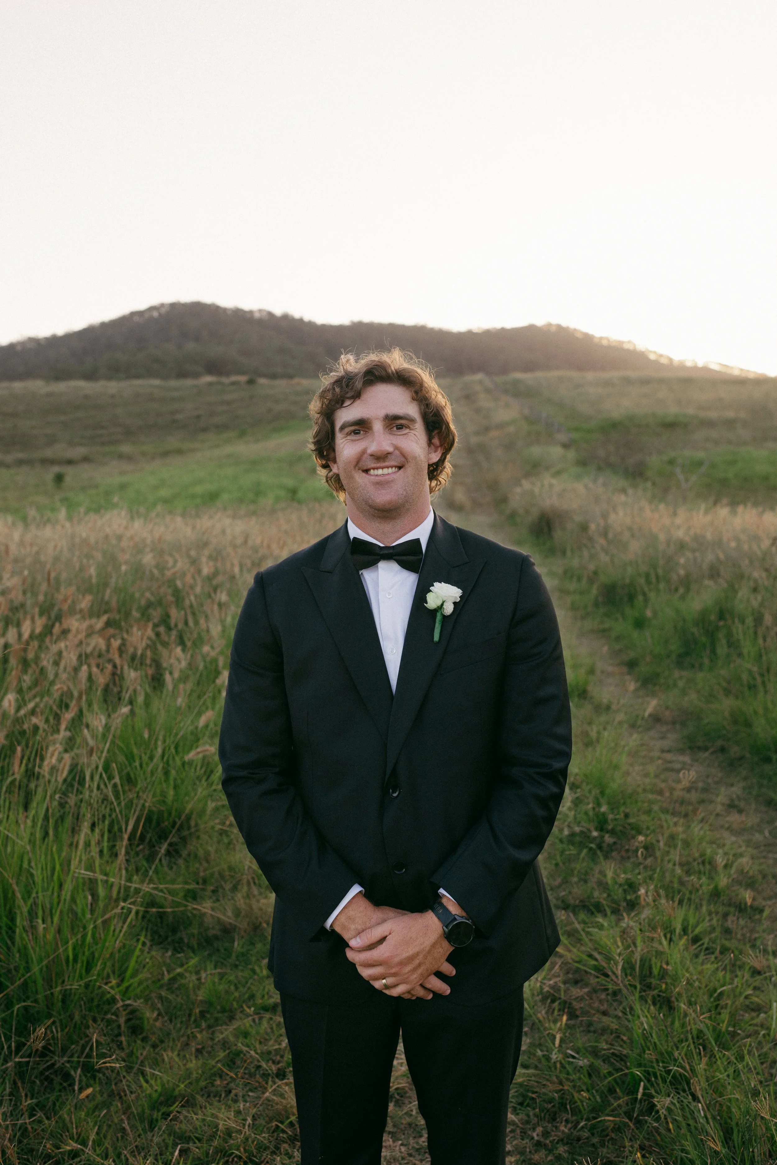 A man dressed in a black tuxedo with a white dress shirt and black bow tie, standing outdoors in a field with green grass and a mountain in the background during sunset. He is smiling and has his hands clasped in front of him.