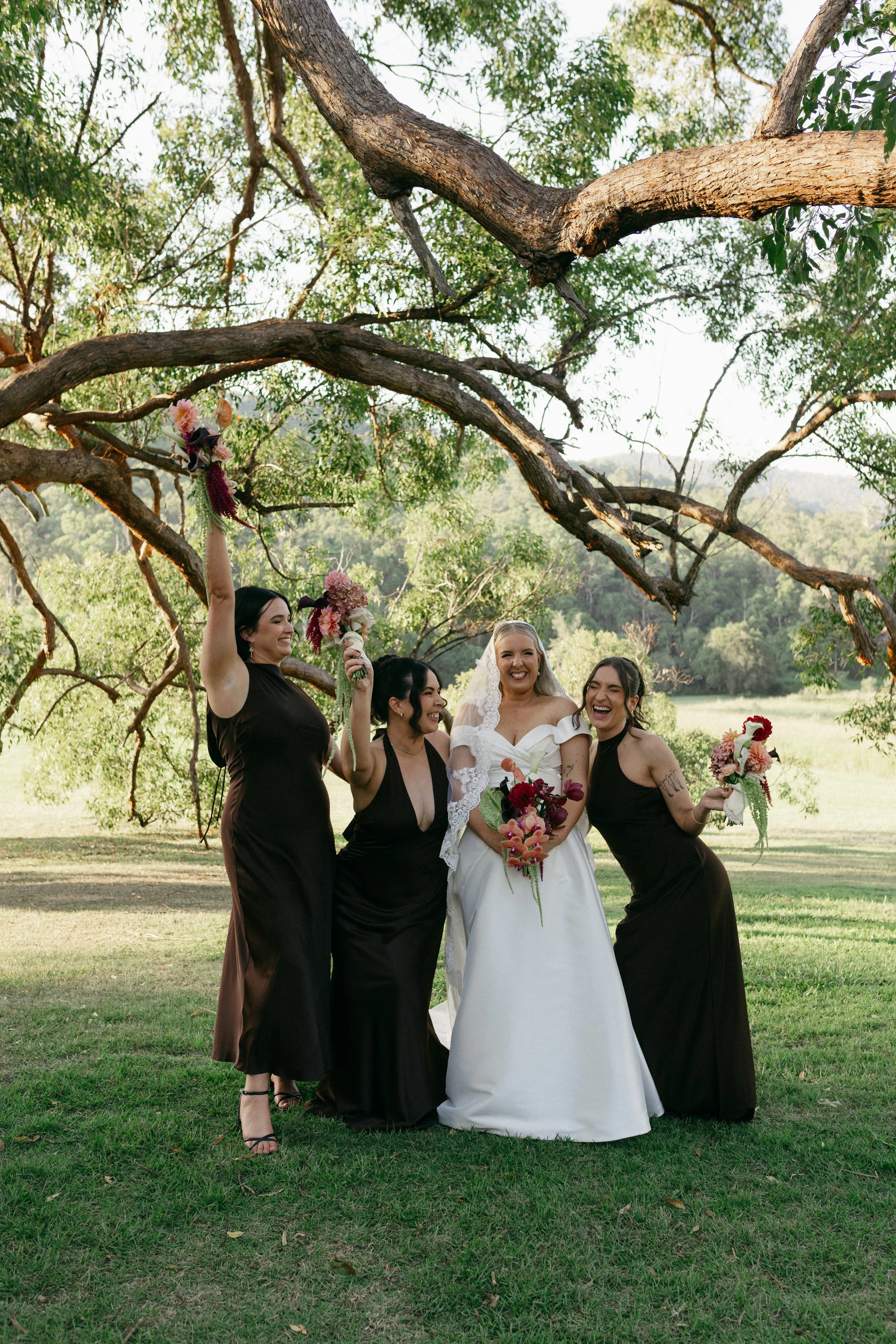 A group of women, including a bride in a white wedding gown, standing outdoors under large tree branches with a green landscape in the background. They are smiling and holding bouquets, celebrating a wedding.