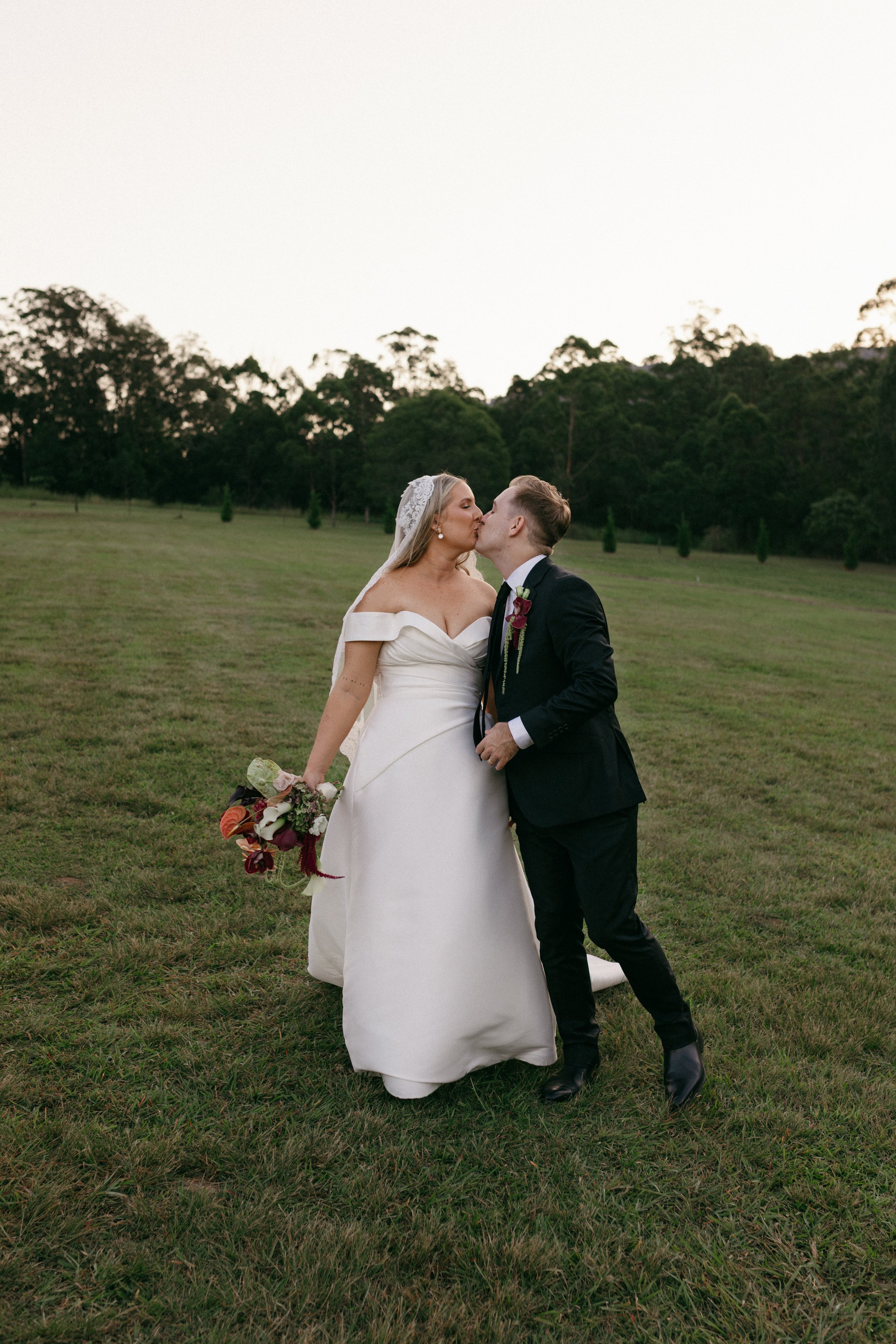A bride and groom kiss outdoors on a grassy field during their wedding, with trees and a clear sky in the background.