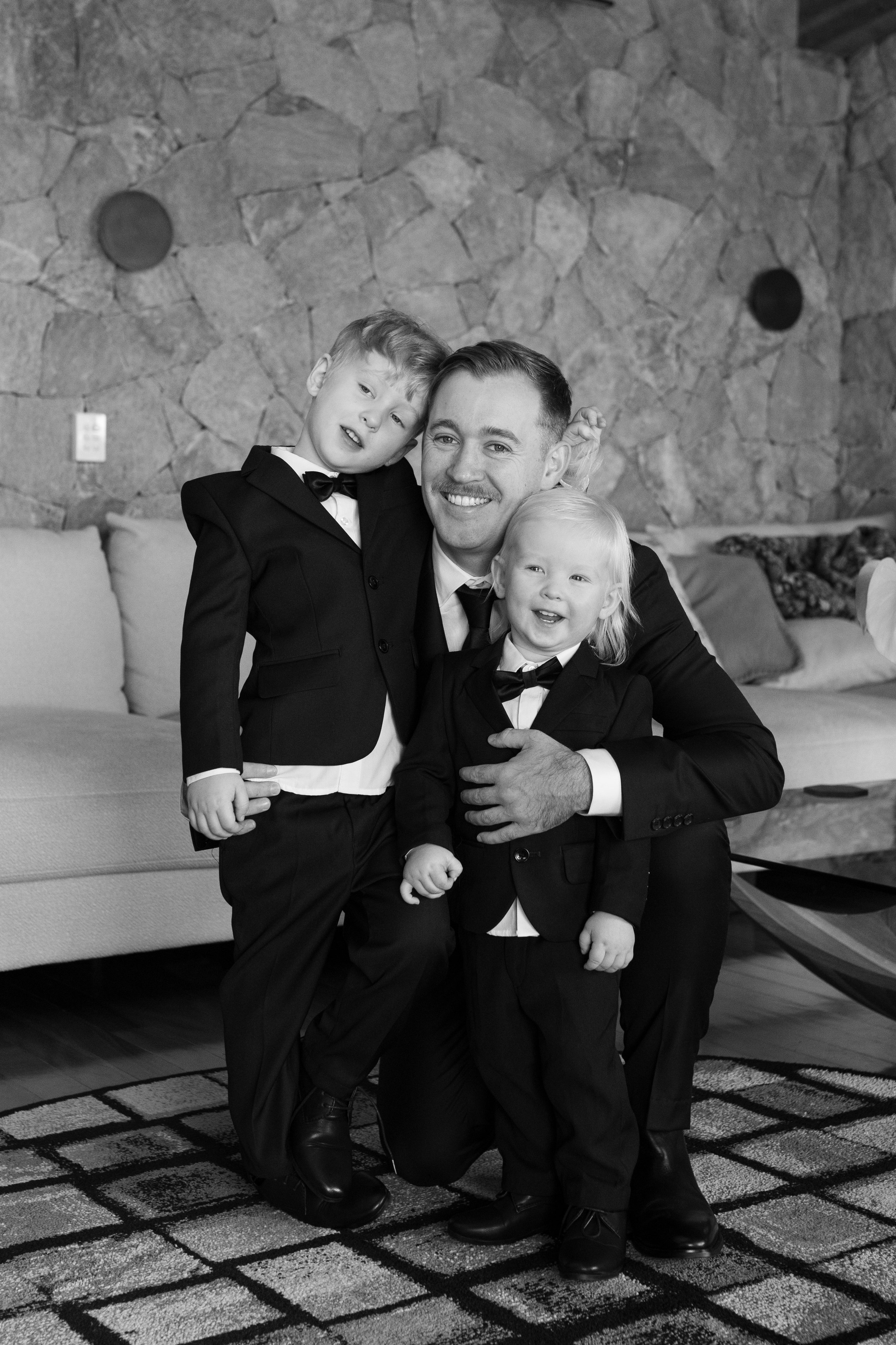 A man in a suit with two children in tuxedos, all smiling and posing together indoors, with a stone wall and patterned carpet in the background.