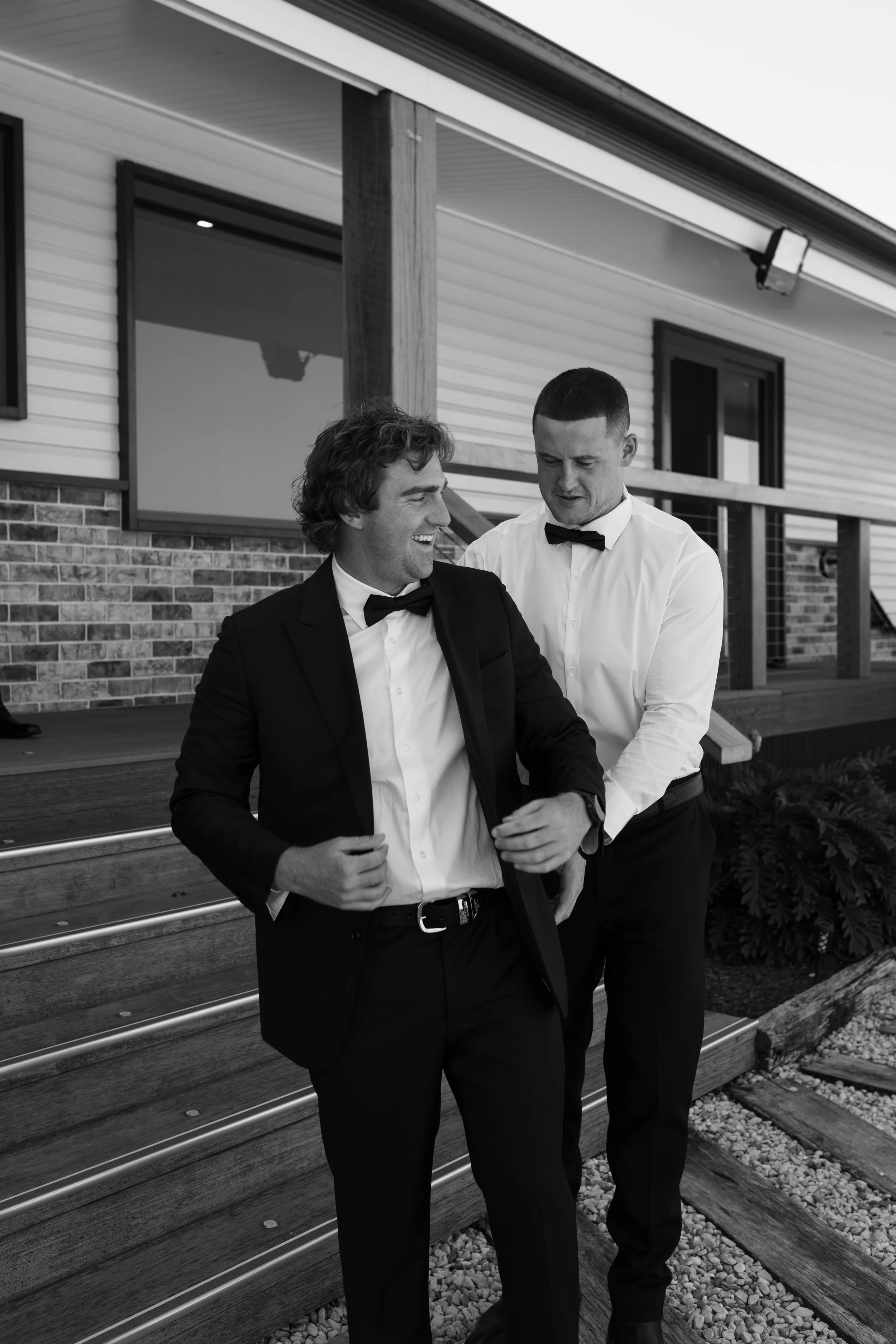 Two men dressed in tuxedos are standing outdoors near a house with siding and windows, engaged in conversation.
