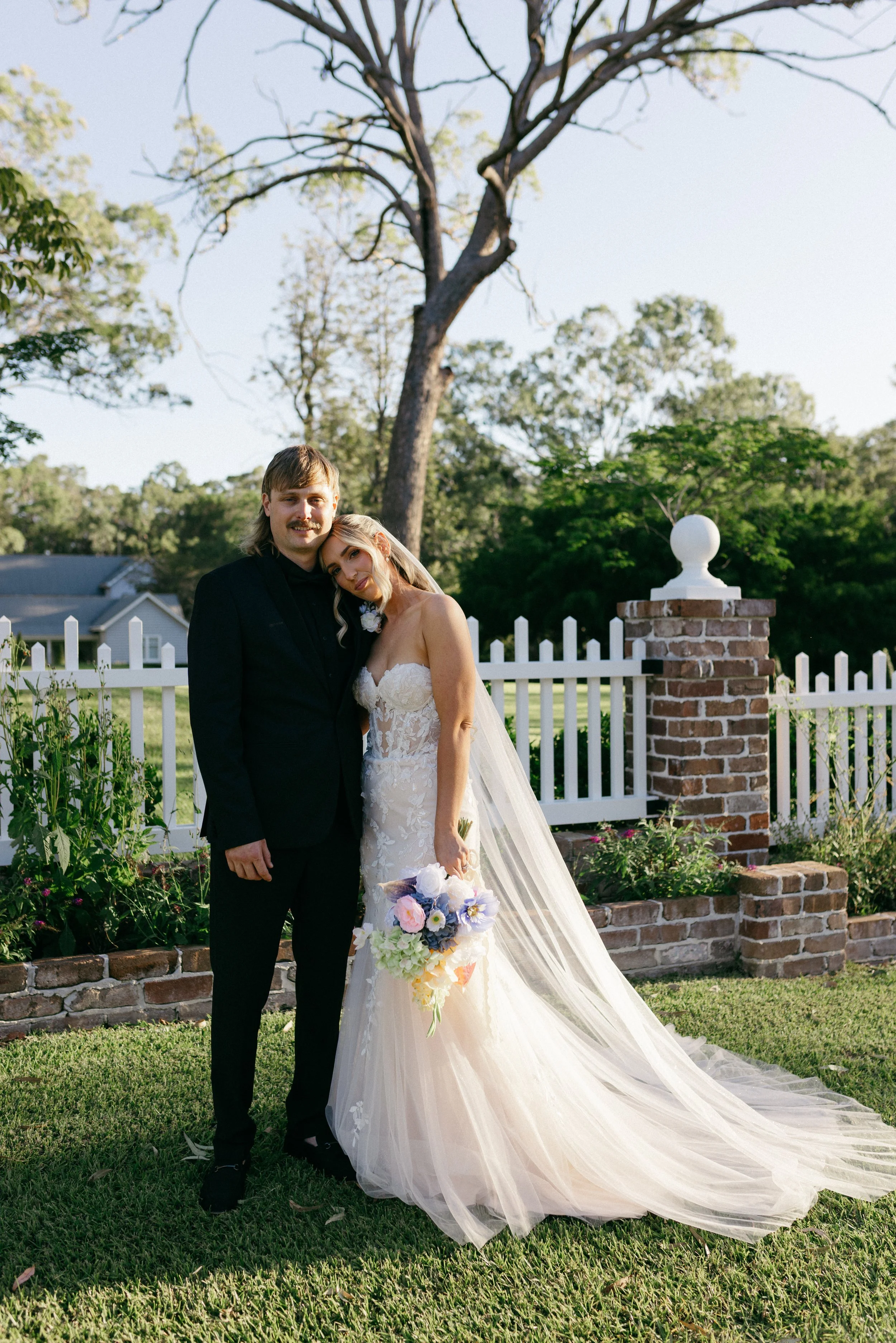 A bride and groom standing close together outdoors, with the bride holding a colorful bouquet of flowers, in front of a white picket fence and brick pillars, beneath a large tree, during daytime.