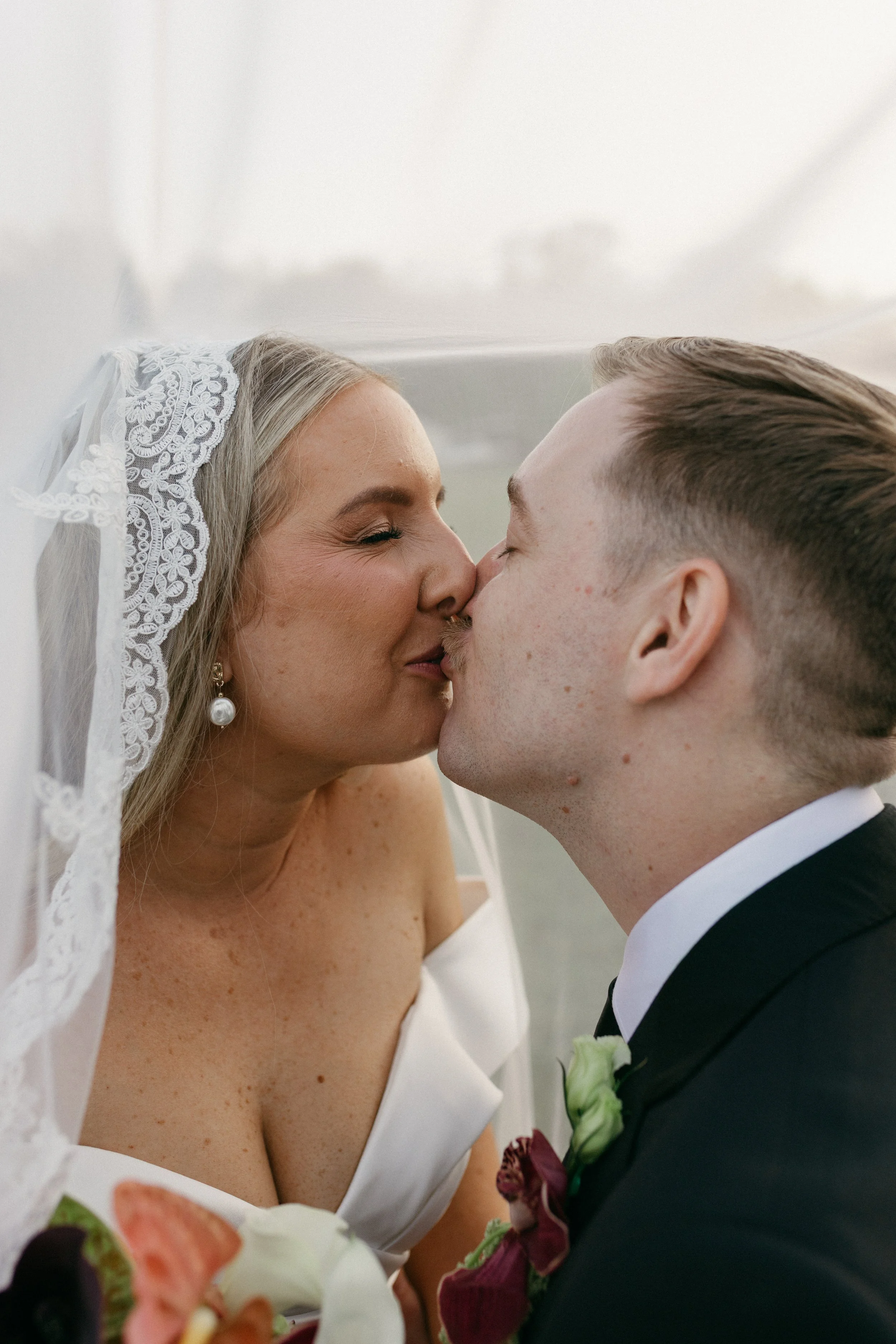 A bride and groom sharing a kiss, close-up shot, bride wearing a white dress and veil with lace trim, groom in a black suit with white shirt and boutonniere.