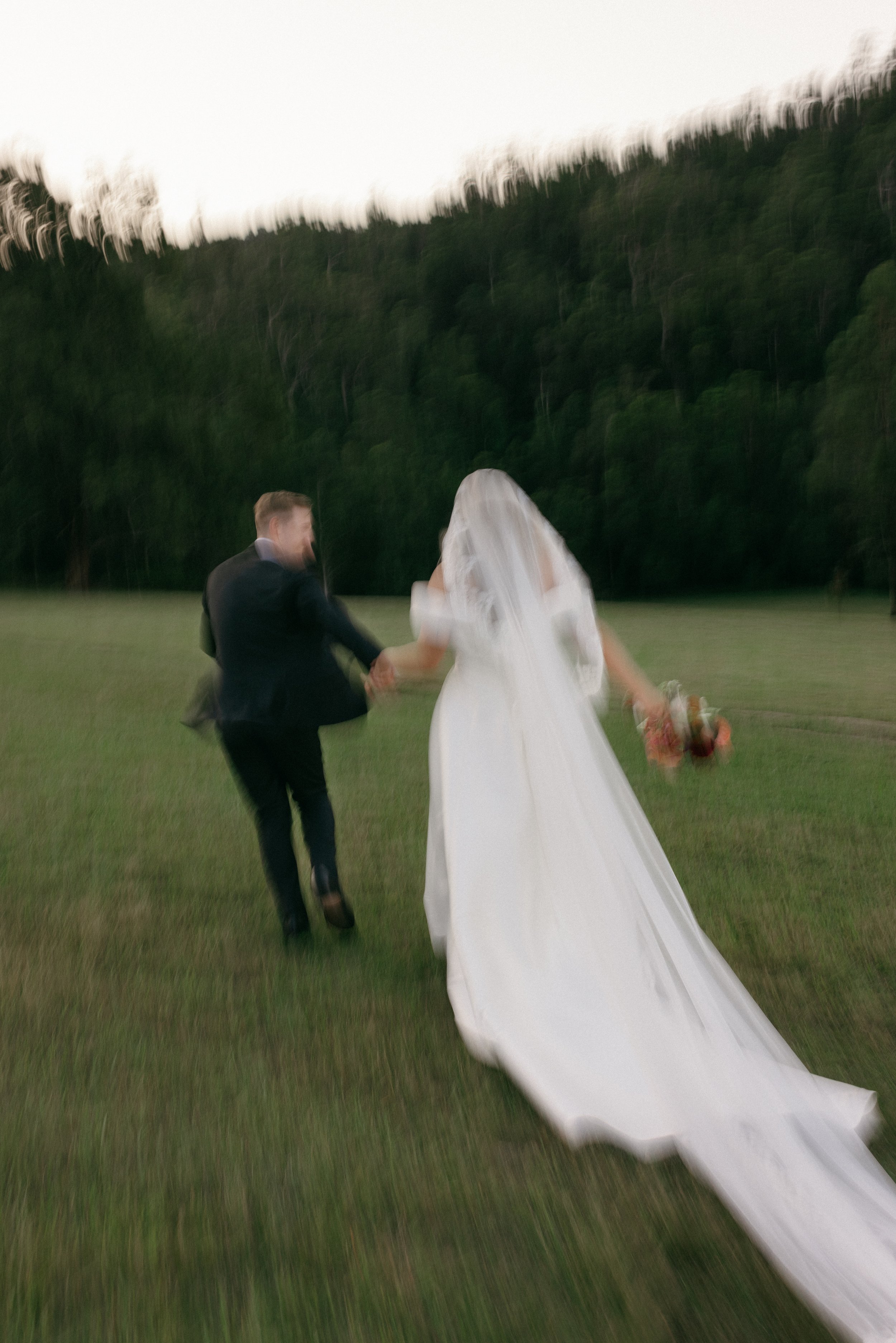 A bride and groom holding hands and walking on a grassy field with a backdrop of trees, with motion blur in the image.