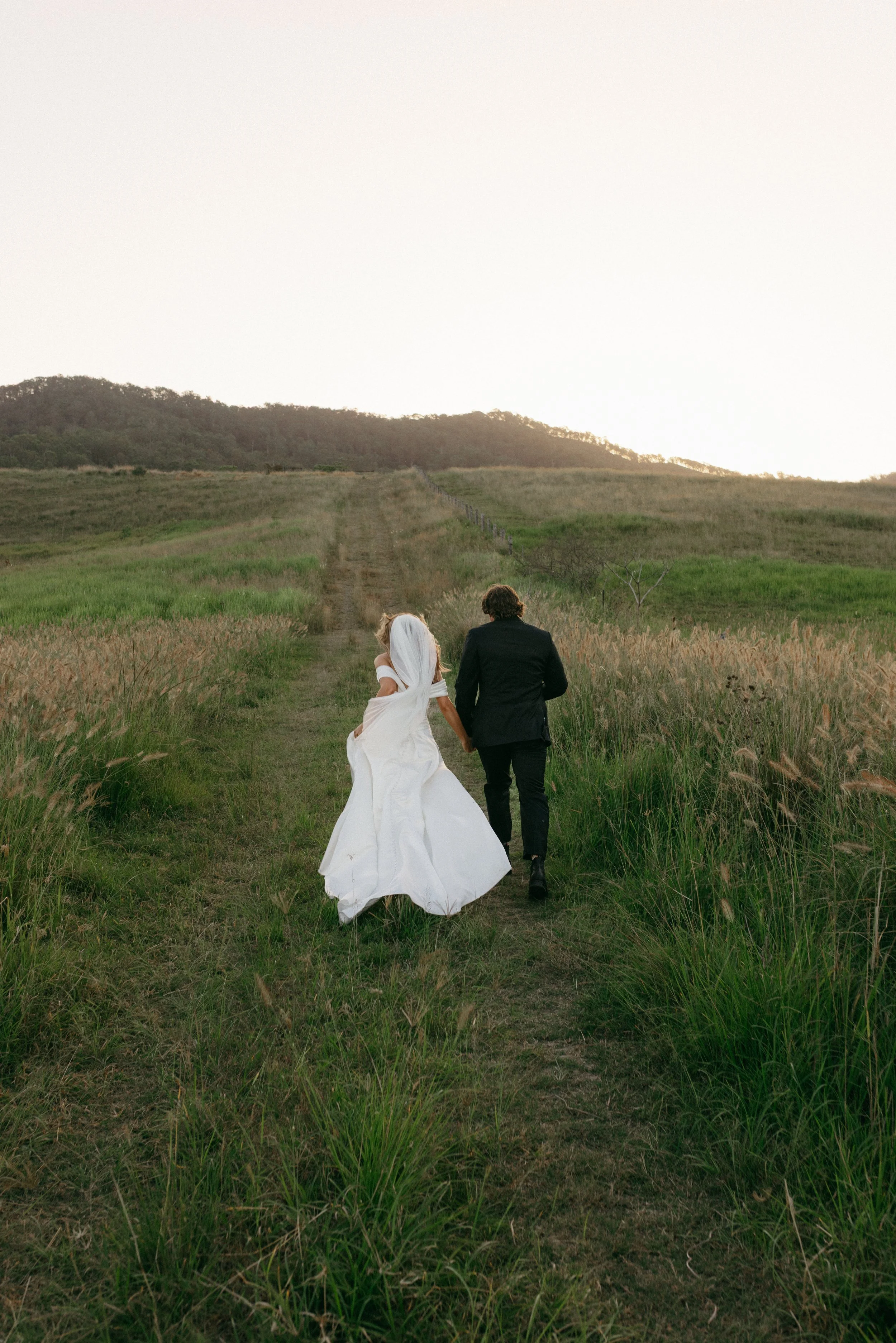 A bride and groom walking hand in hand down a dirt rural path through tall grass, with hills in the background at sunset.
