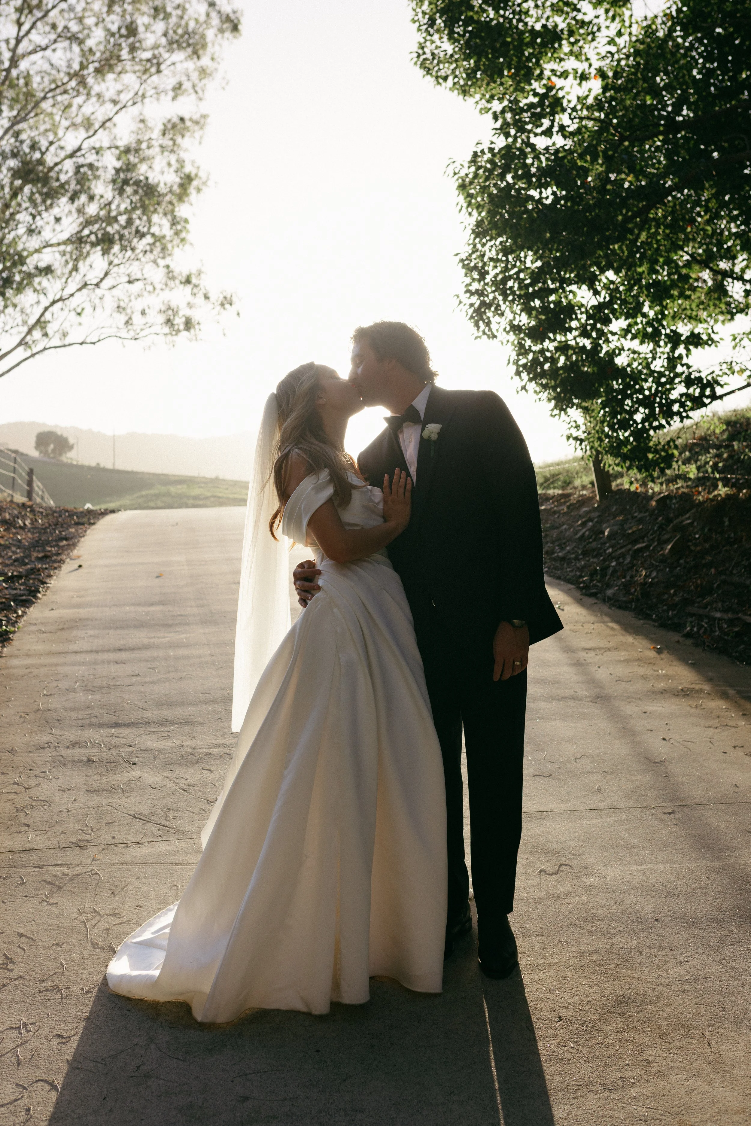 A bride and groom kiss outdoors during sunset; the bride is in a white wedding dress with a veil, and the groom is in a black tuxedo with a bow tie. They are on a paved road lined with trees.
