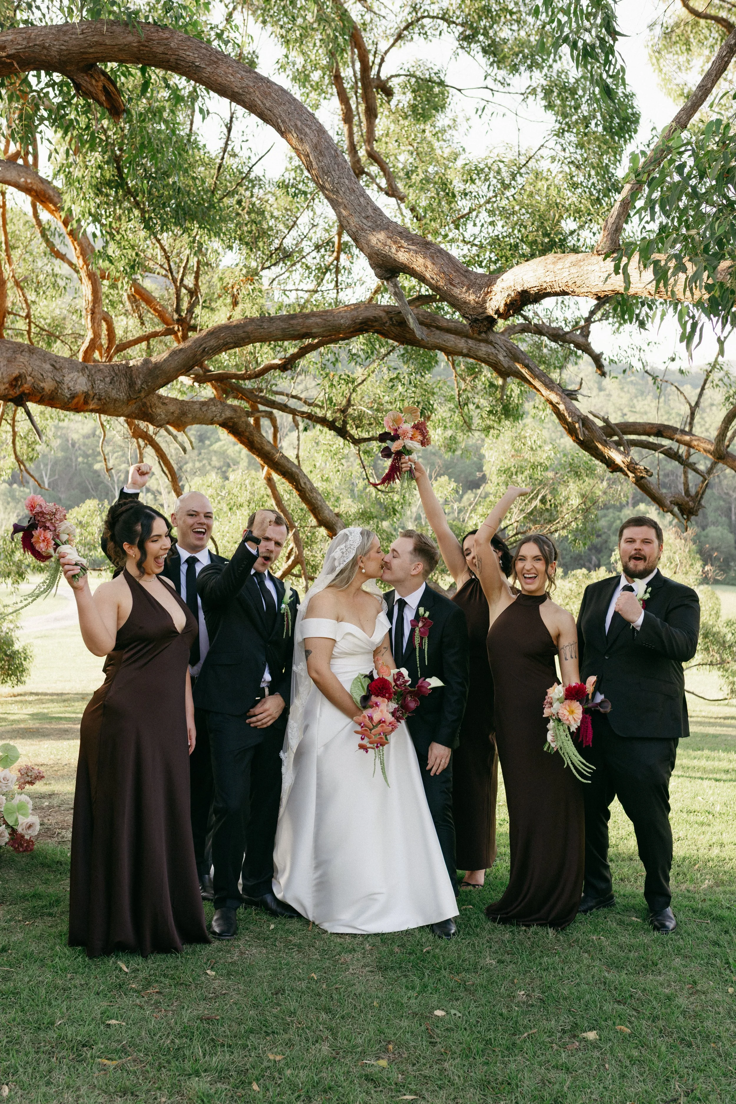 A group of people dressed in formal attire celebrating a wedding outdoors under a large tree, with some holding bouquets and raising their fists or hands in celebration.