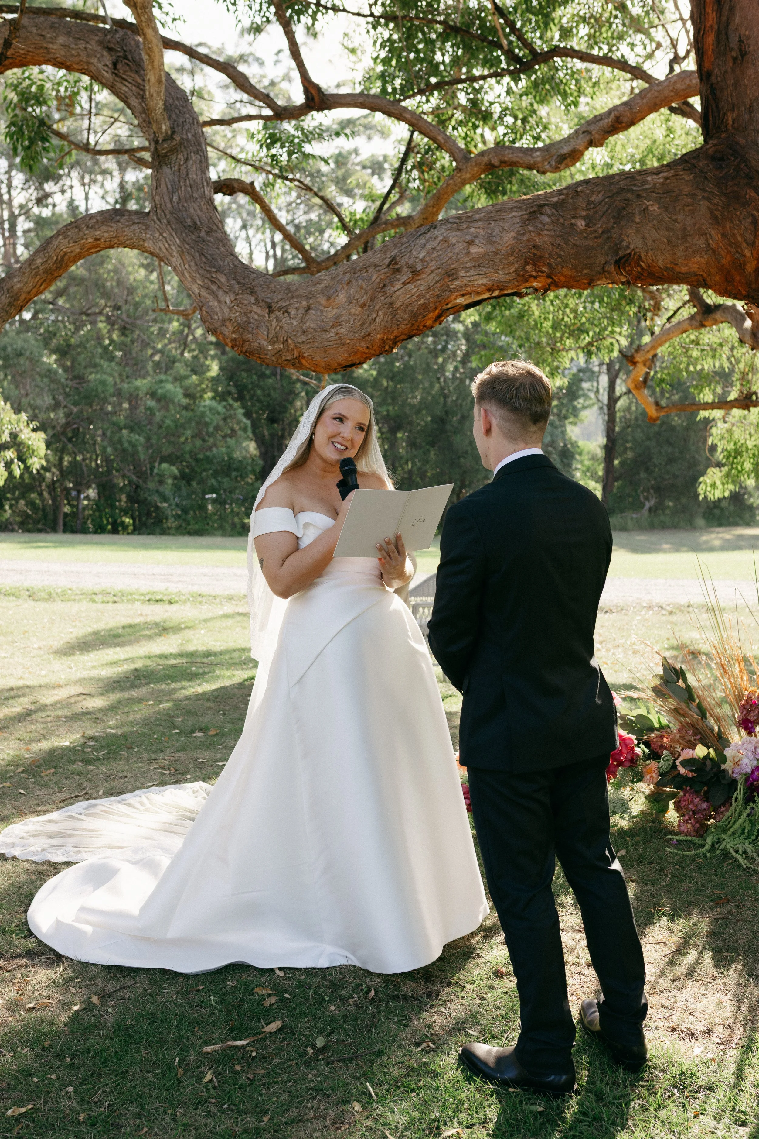 A bride in a white wedding dress holding a book and speaking into a microphone, with a groom in a black suit facing her, outdoors in a garden with a large tree overhead and flowers nearby.