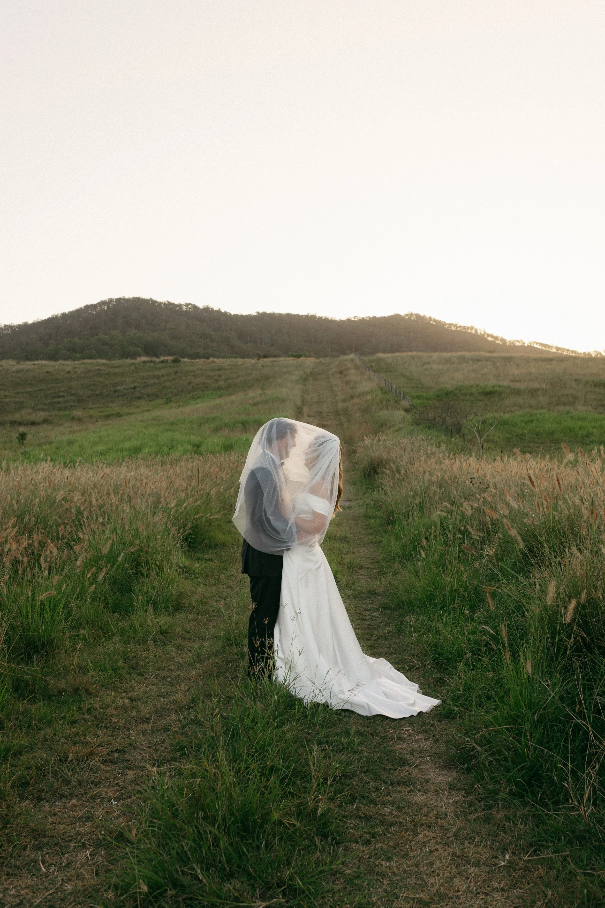 A couple dressed in wedding attire standing on a grassy path in a rural landscape, with green fields and hills in the background, and a cloudy sky overhead.