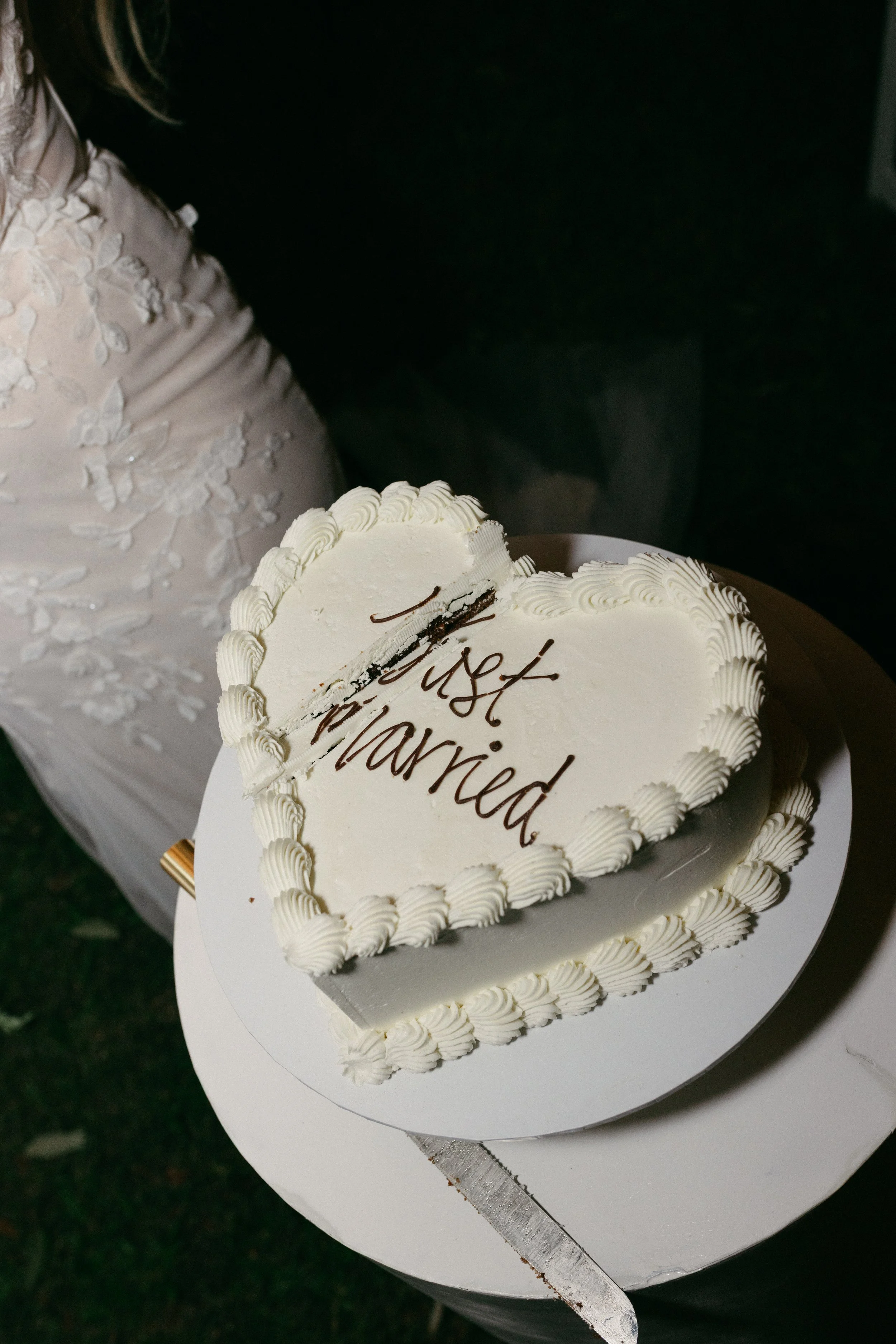 Heart-shaped wedding cake with white frosting and decorative piping, with a message written in chocolate on top. Part of a person in a lace wedding dress is visible in the background.