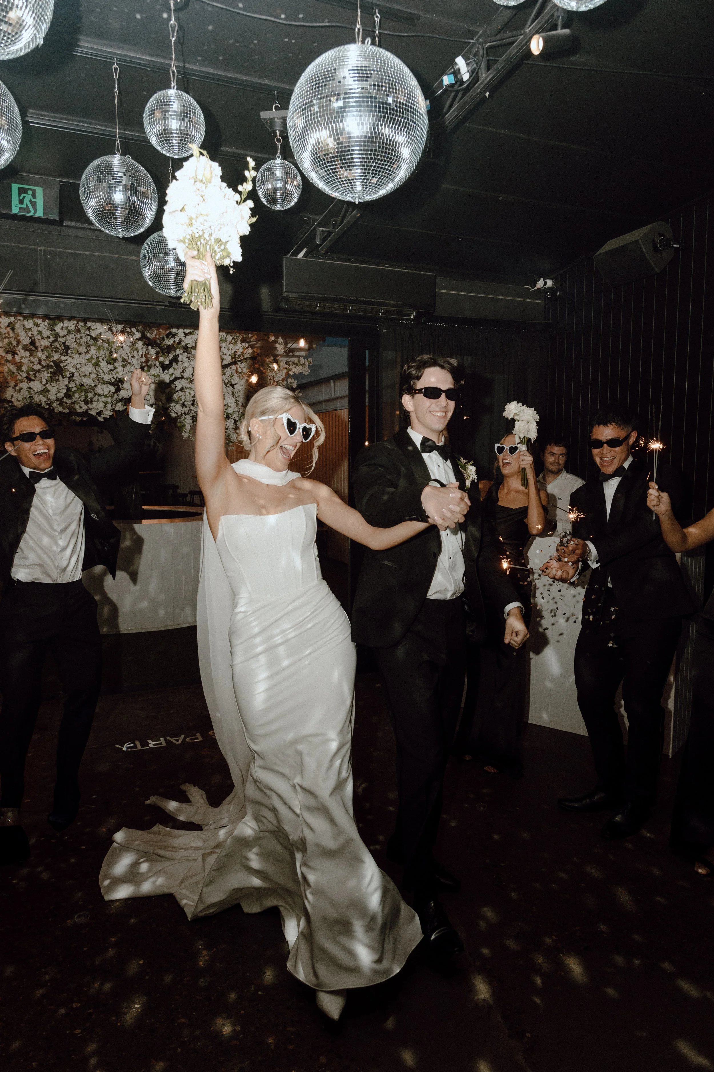 People celebrating at a party, with one woman in a white dress holding a bouquet of flowers and wearing heart-shaped sunglasses, and others in tuxedos and dresses holding sparklers. There are disco balls hanging from the ceiling.