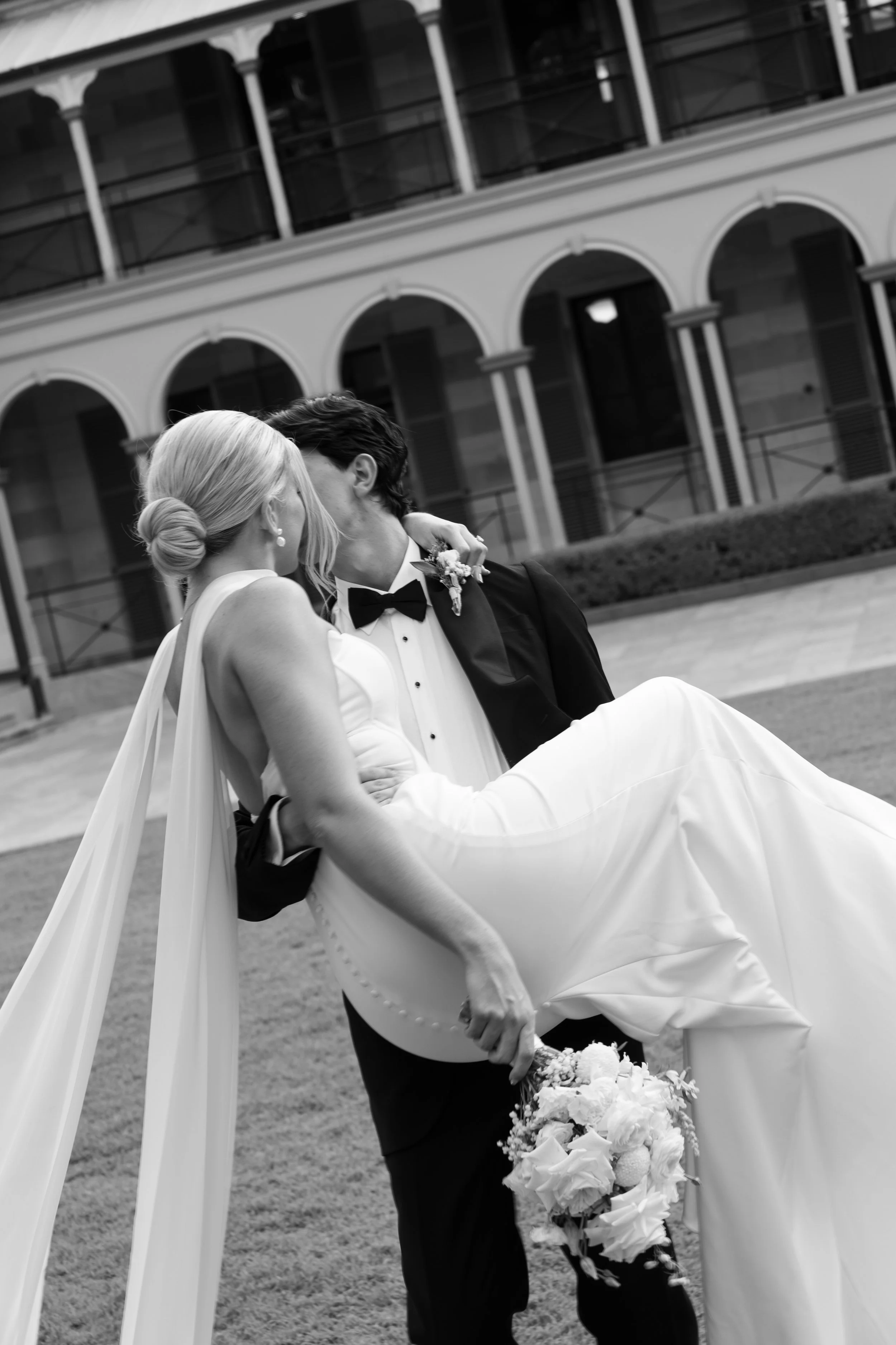 A black-and-white photo of a bride and groom kissing outside a building with arches and balconies. The bride has platinum blonde hair styled in a bun and is wearing pearl earrings and a strapless wedding dress. The groom has dark hair and is dressed 