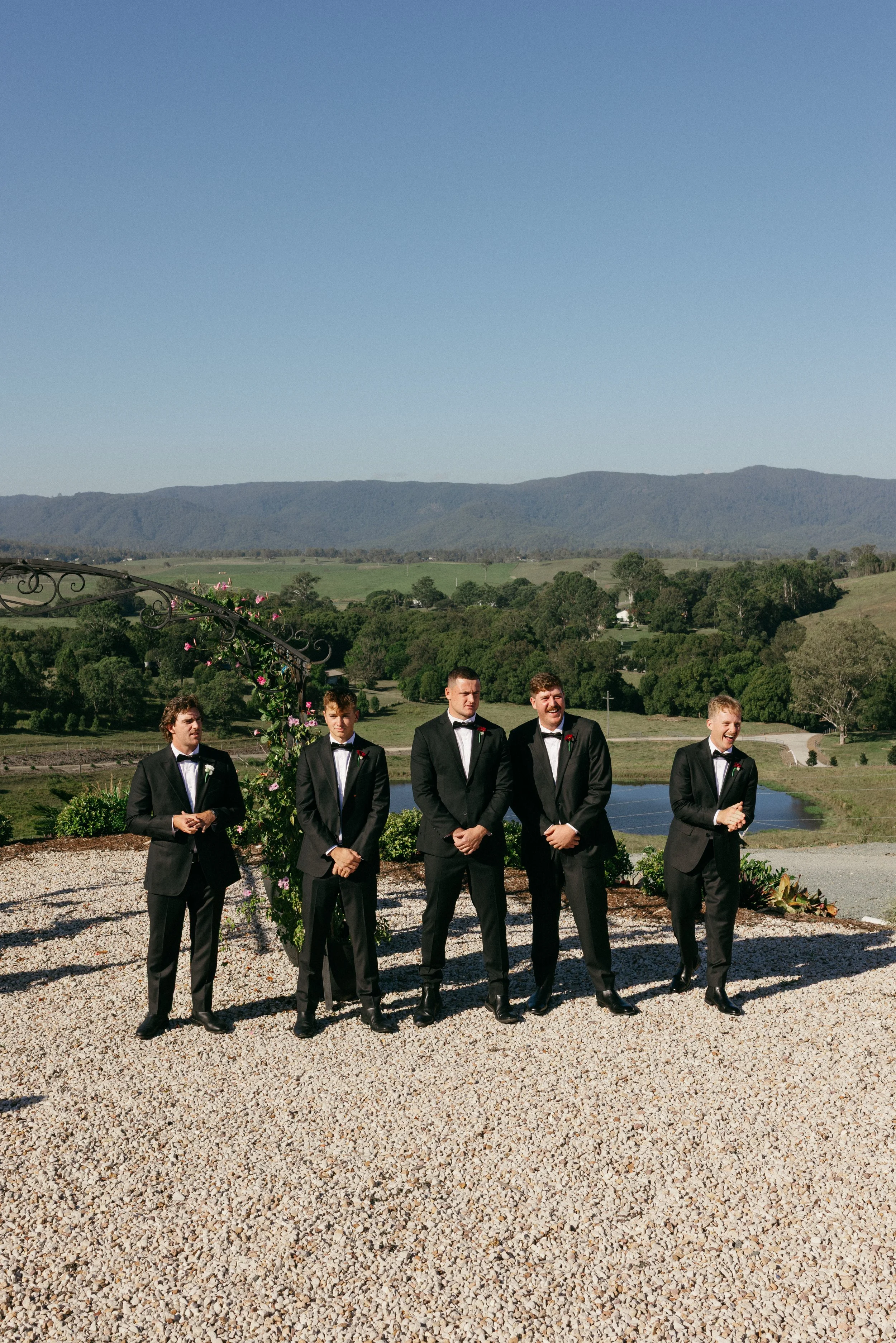 Group of five men dressed in black tuxedos standing outdoors on a gravel surface, with mountains and a clear blue sky in the background, during a wedding ceremony.