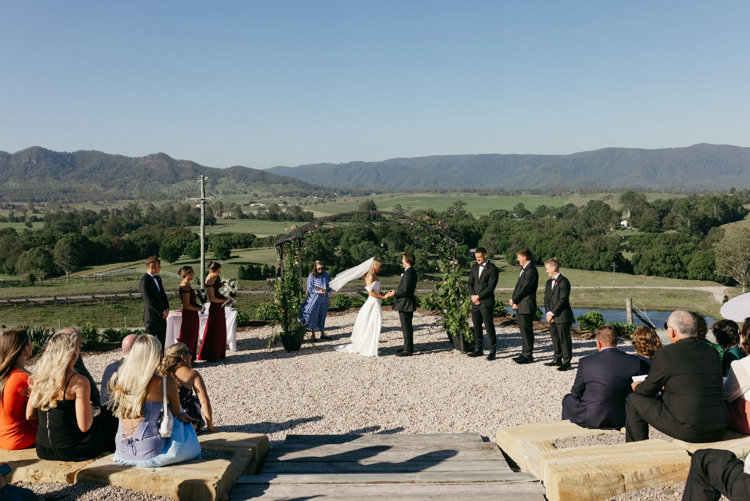 Wedding ceremony outdoors with couples exchanging vows under a floral arch, surrounded by seated guests, with scenic mountains and clear blue sky in the background.