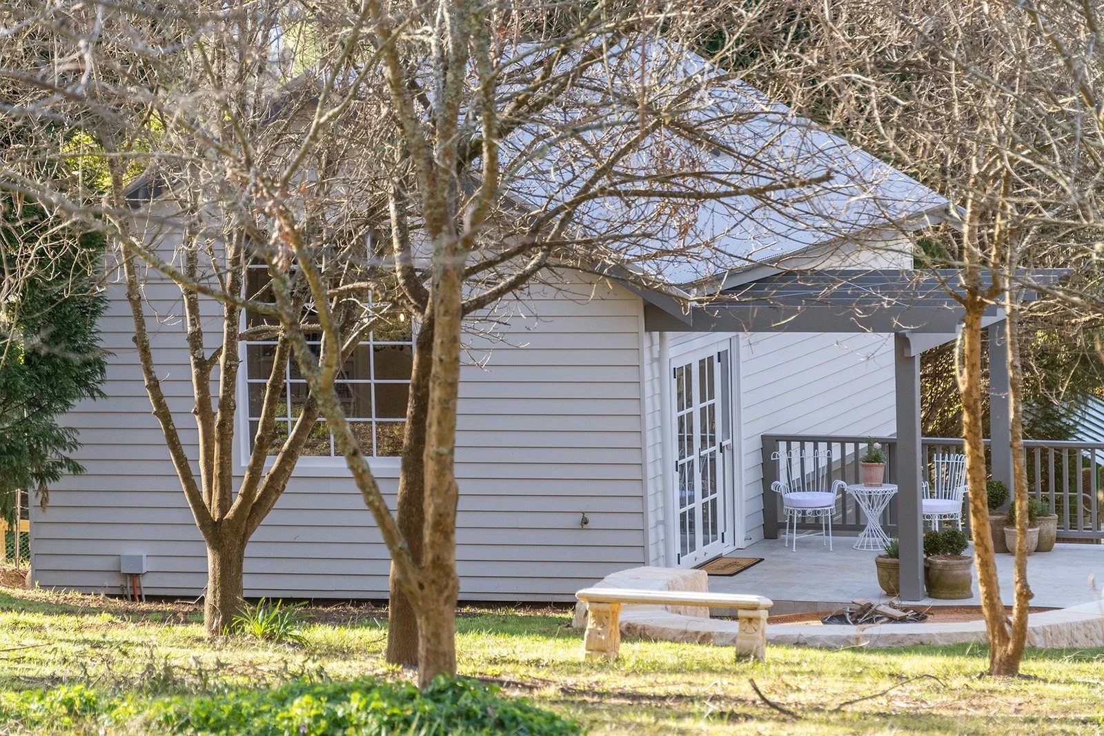 A house with a patio area, white siding, and a gray roof, viewed through leafless trees in a yard with grass and potted plants.