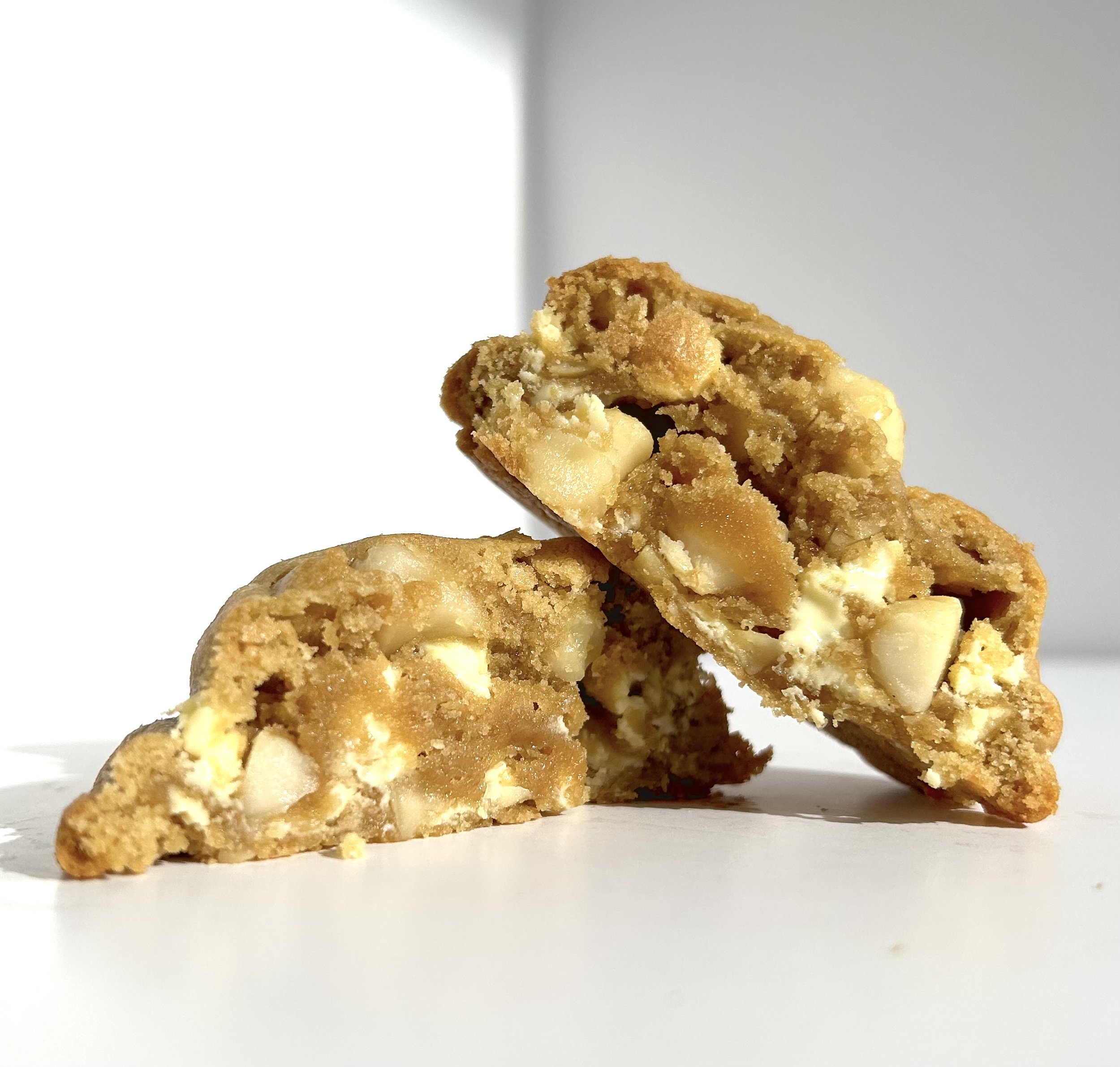 Close-up of two chocolate chip cookies broken in half, with white chocolate chips visible, on a white surface against a blurred light background.